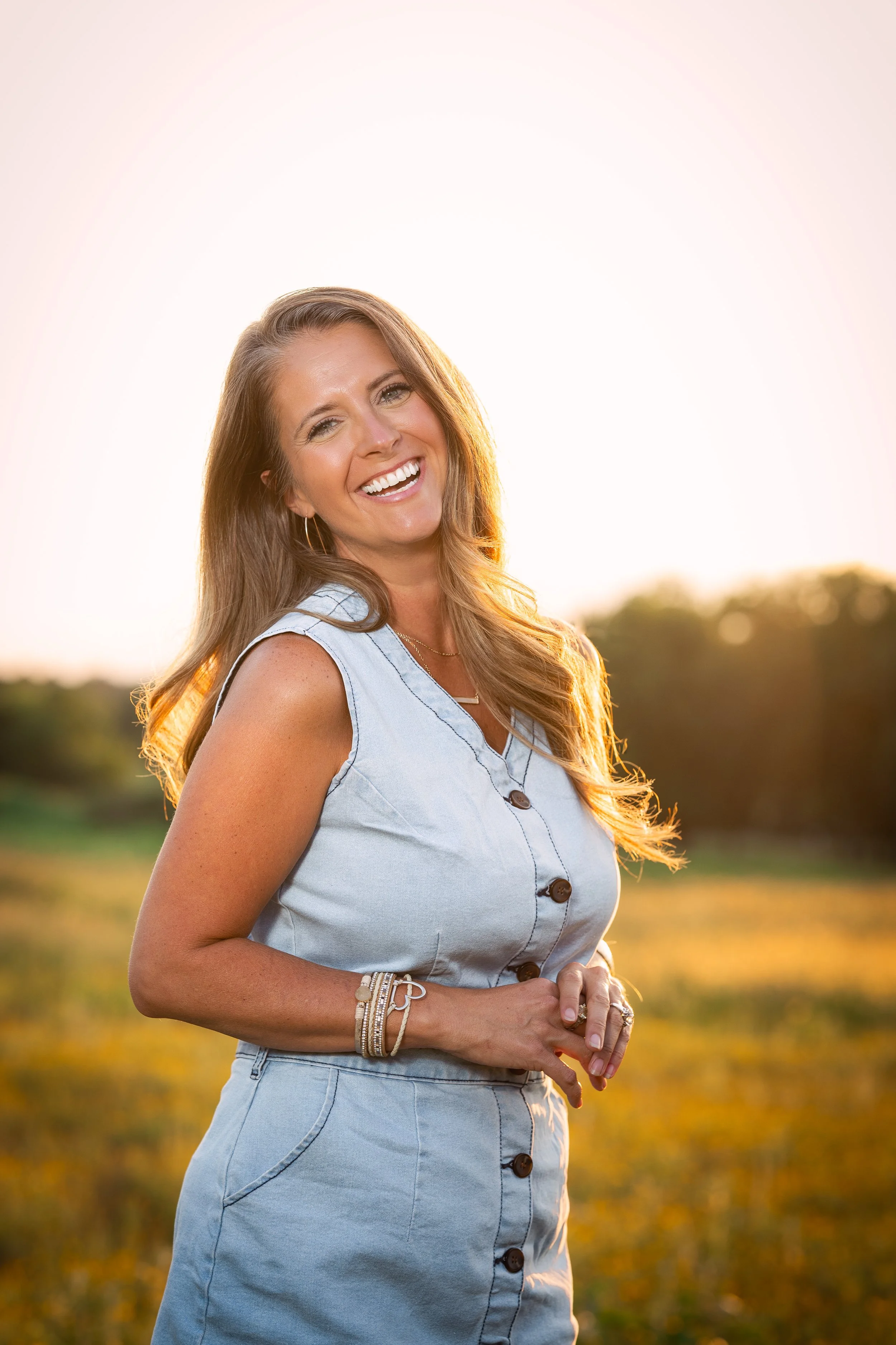 Woman smiling outdoors during sunset, wearing a sleeveless denim top, matching shorts, and jewelry.