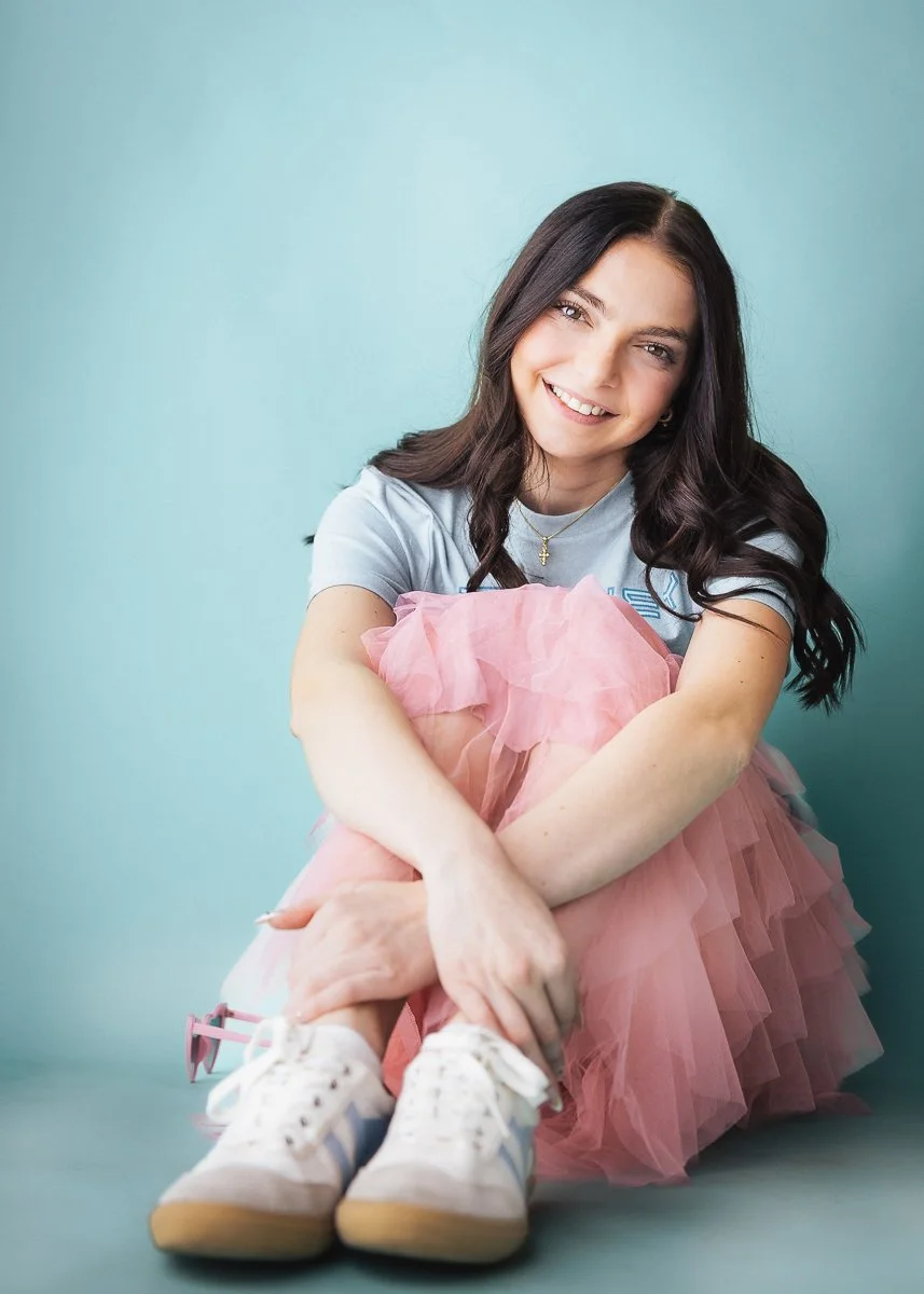A young woman with long dark hair sitting on the floor against a light blue background, smiling, wearing a light gray t-shirt, a pink tutu skirt, white sneakers, and holding pink sunglasses.