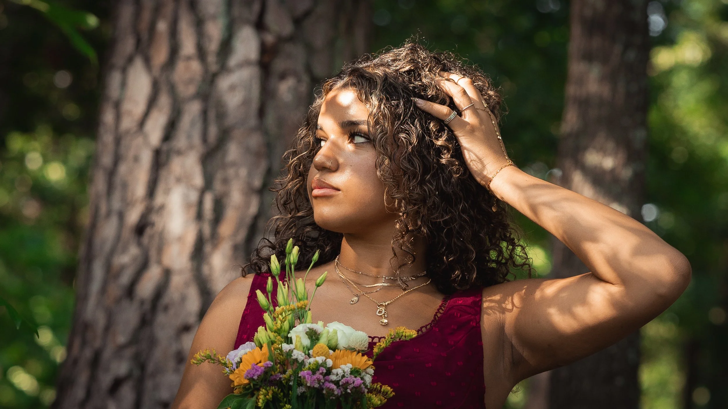A woman with curly brown hair wearing a sleeveless red dress, holding a bouquet of colorful flowers, standing outdoors in a forest near a tree, with sunlight filtering through the leaves.