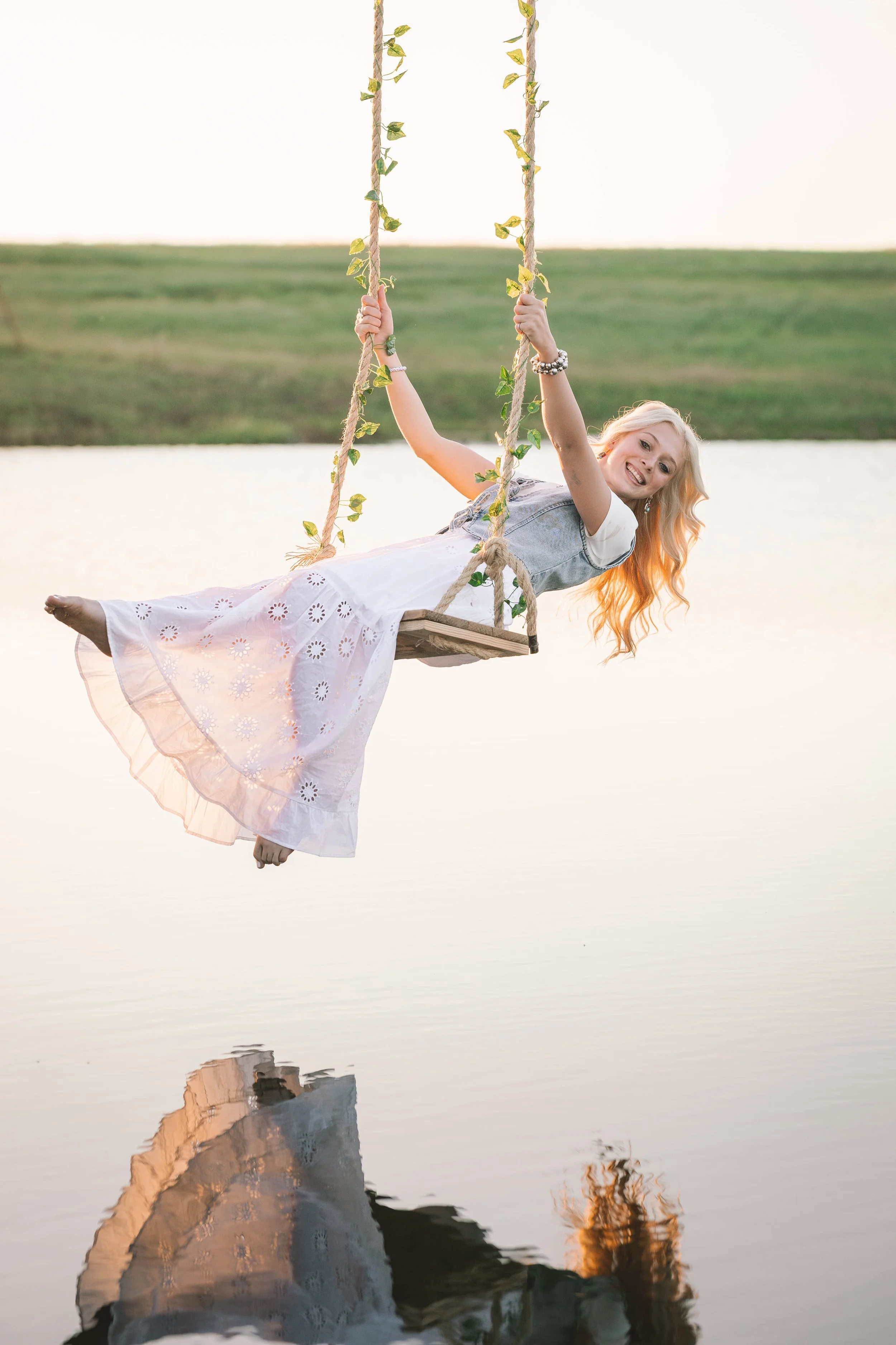 Young girl with long blonde hair wearing a white dress and jean vest swinging on a rope swing over water, smiling at the camera during sunset.
