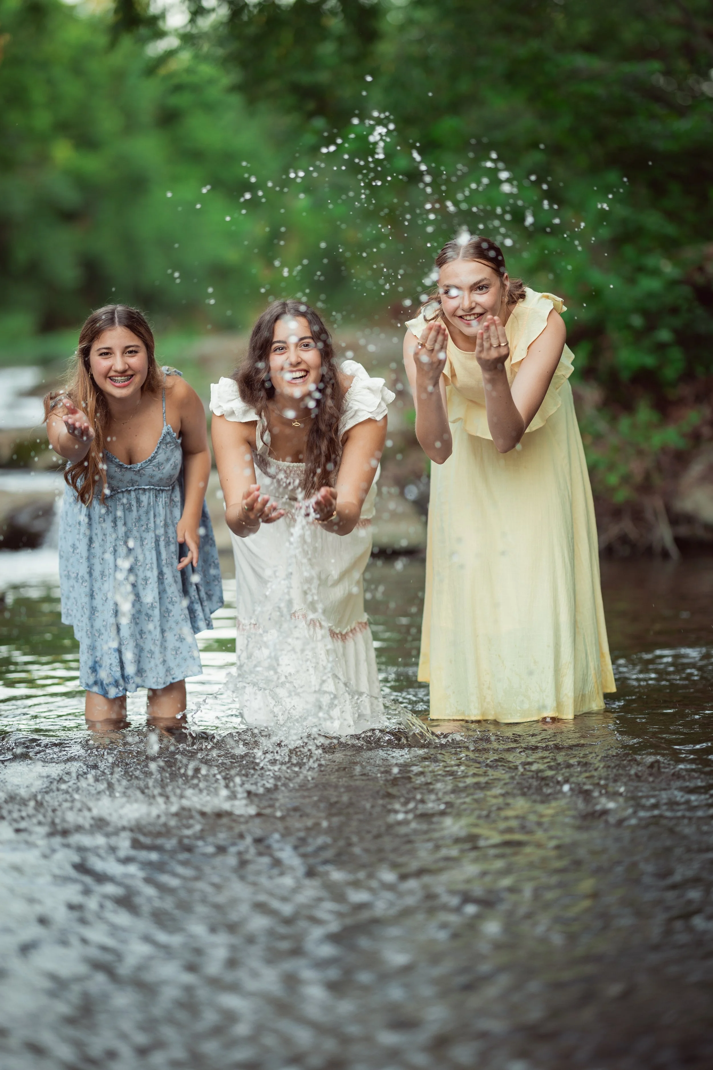 Three young women playing in a creek, splashing water and smiling, surrounded by greenery.