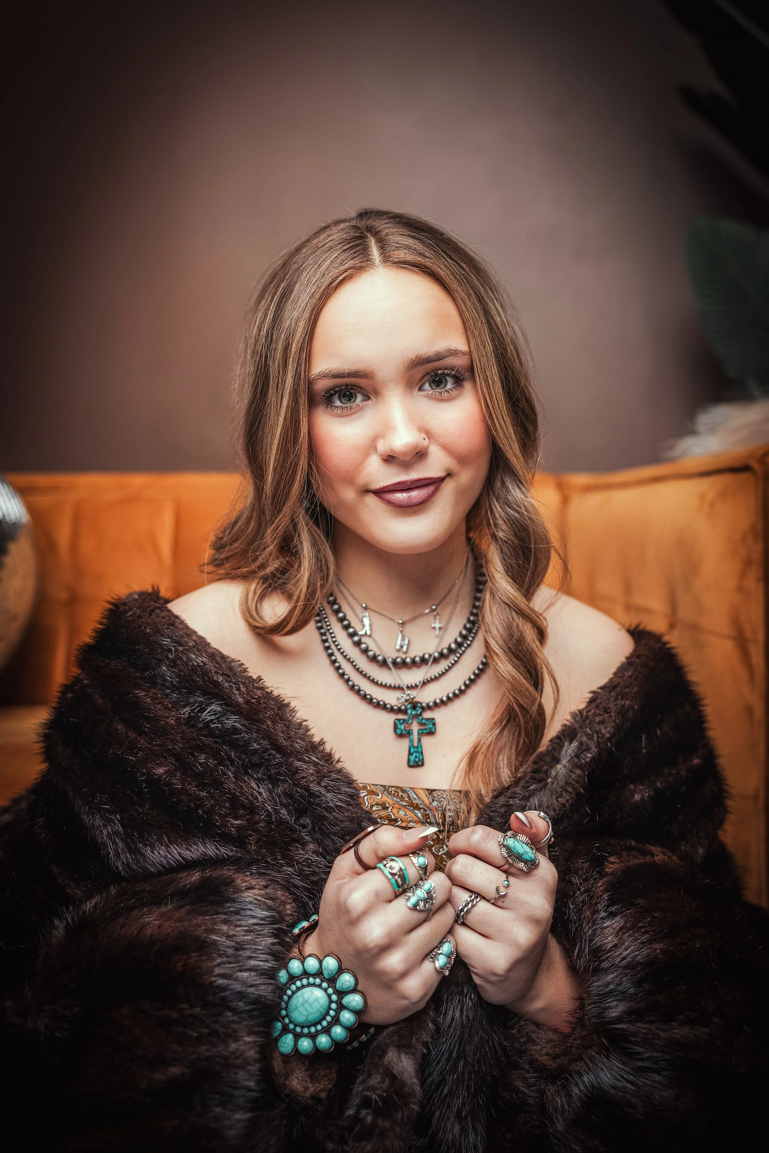 A young woman with shoulder-length light brown hair, wearing multiple layered necklaces, a large turquoise cross pendant, and rings and bracelets with turquoise stones, seated against a brown background with a tufted orange upholstery behind her.