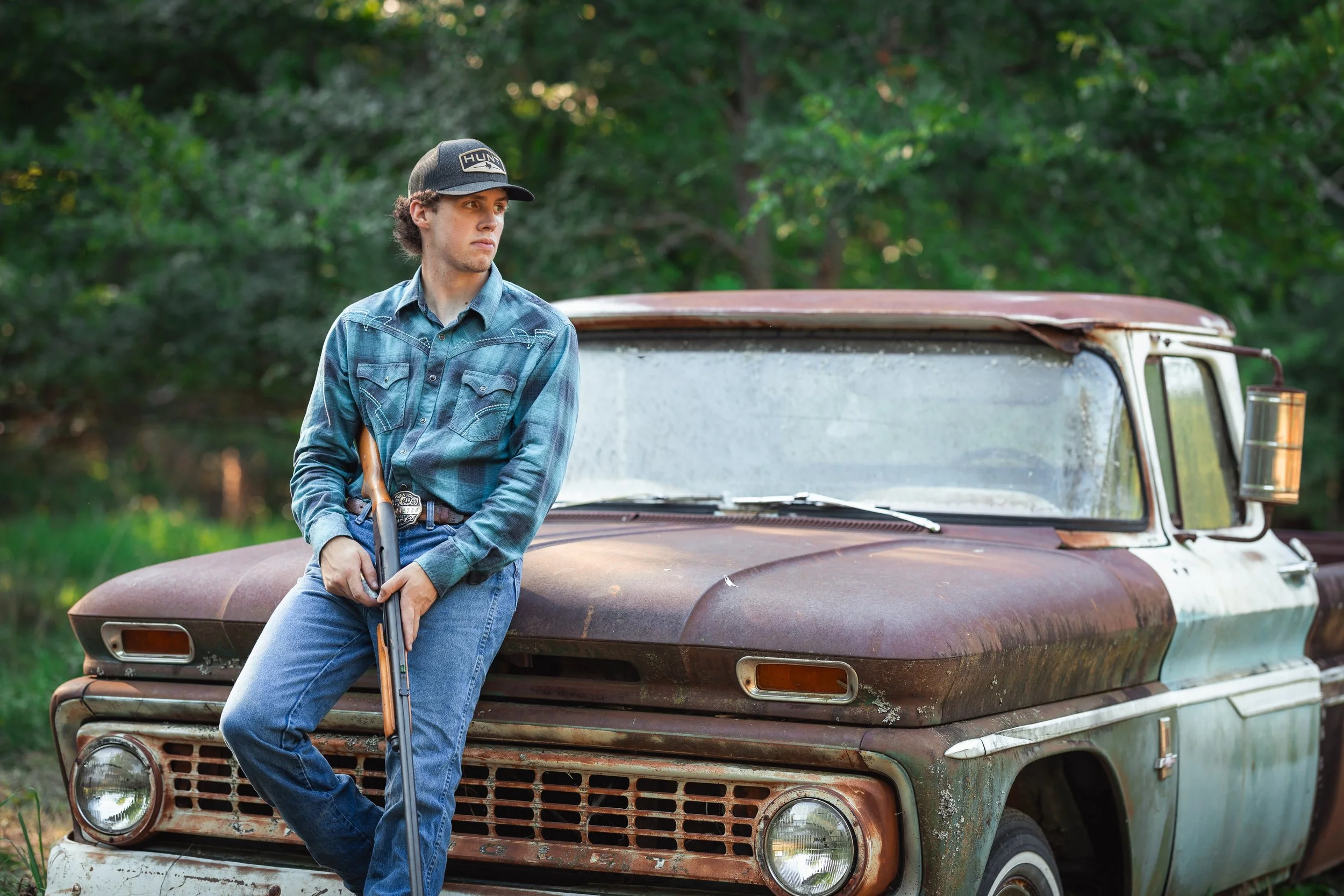 A young man leaning against a rusty, old pickup truck in a wooded outdoor setting, holding a shotgun.
