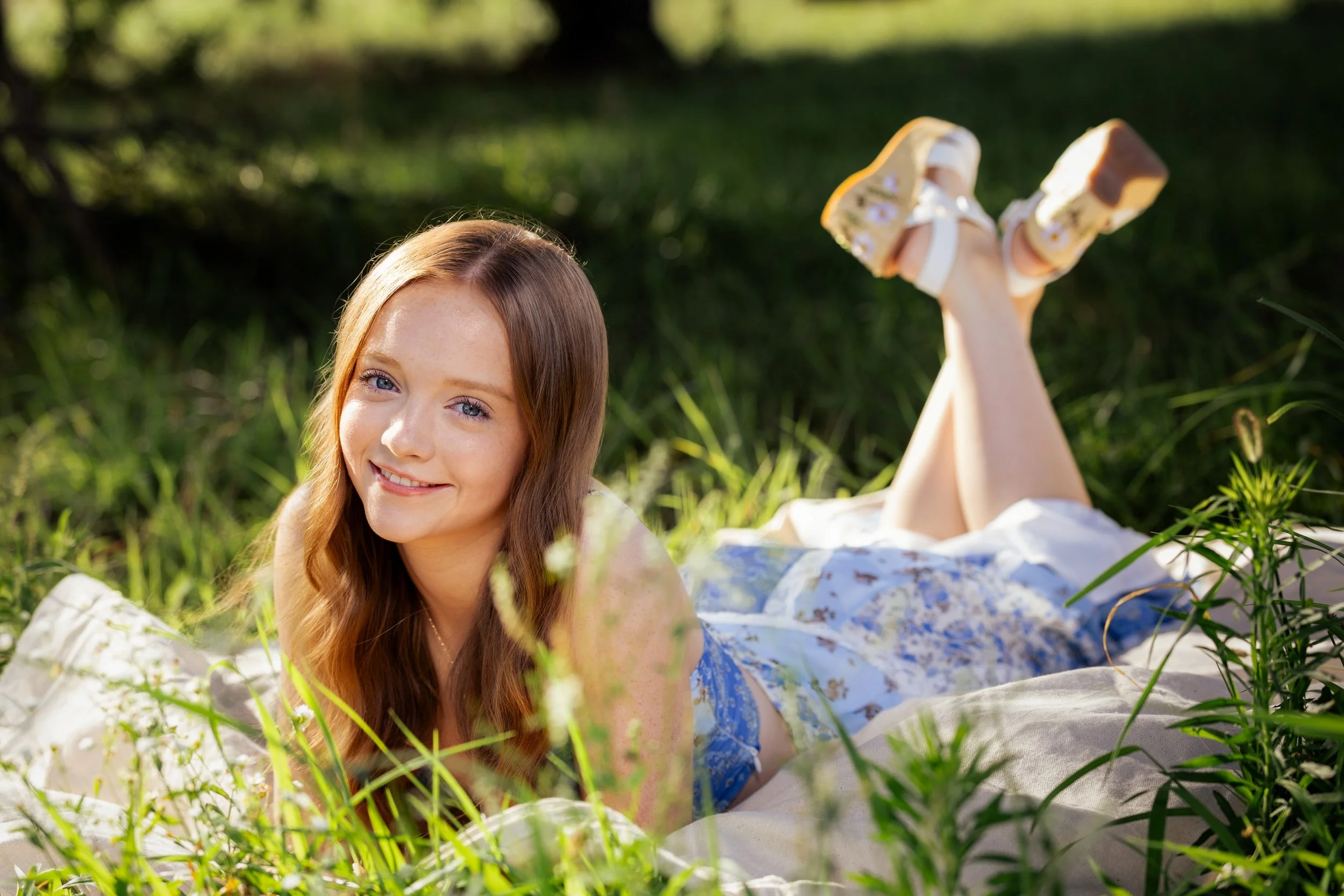 Young girl with long red hair lying on her stomach in grass, smiling at the camera, with her legs raised behind her, wearing yellow shoes.