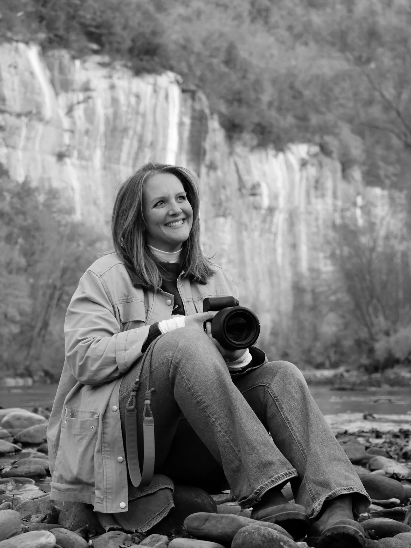 A woman sitting on a rocky riverbank holding a camera, smiling, with a waterfall and trees in the background.