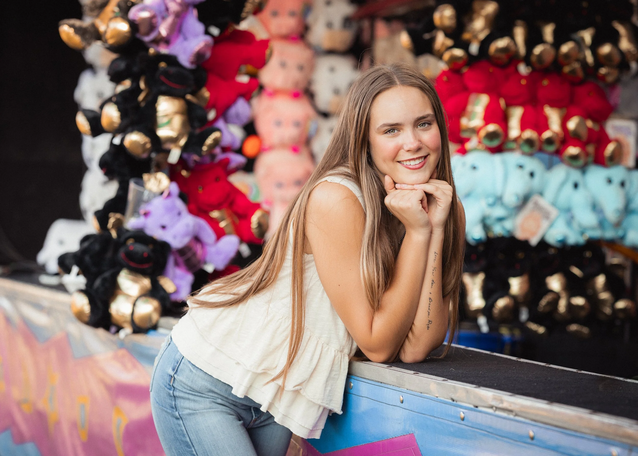 A young woman with long light brown hair smiling at the camera, resting her chin on her hands, while leaning on a carnival game table with plush toys in the background.
