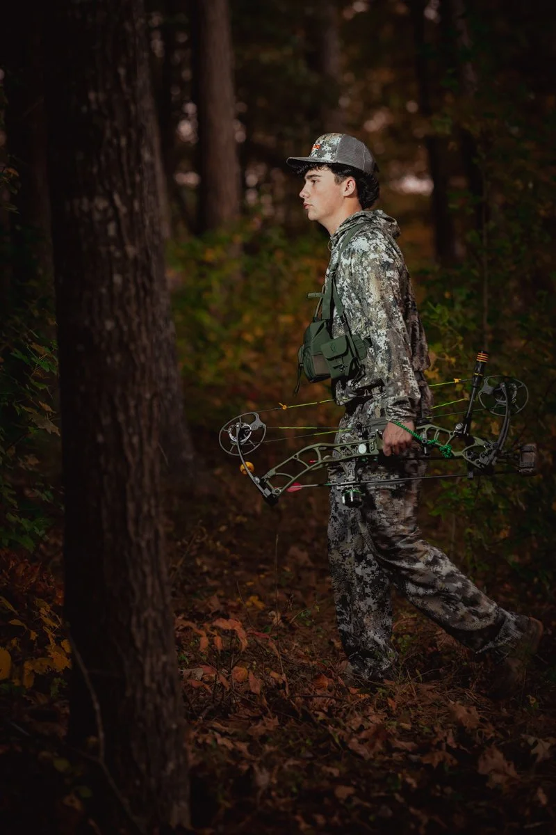 A young man dressed in camouflage clothing standing in a forest, holding a compound bow in his right hand.