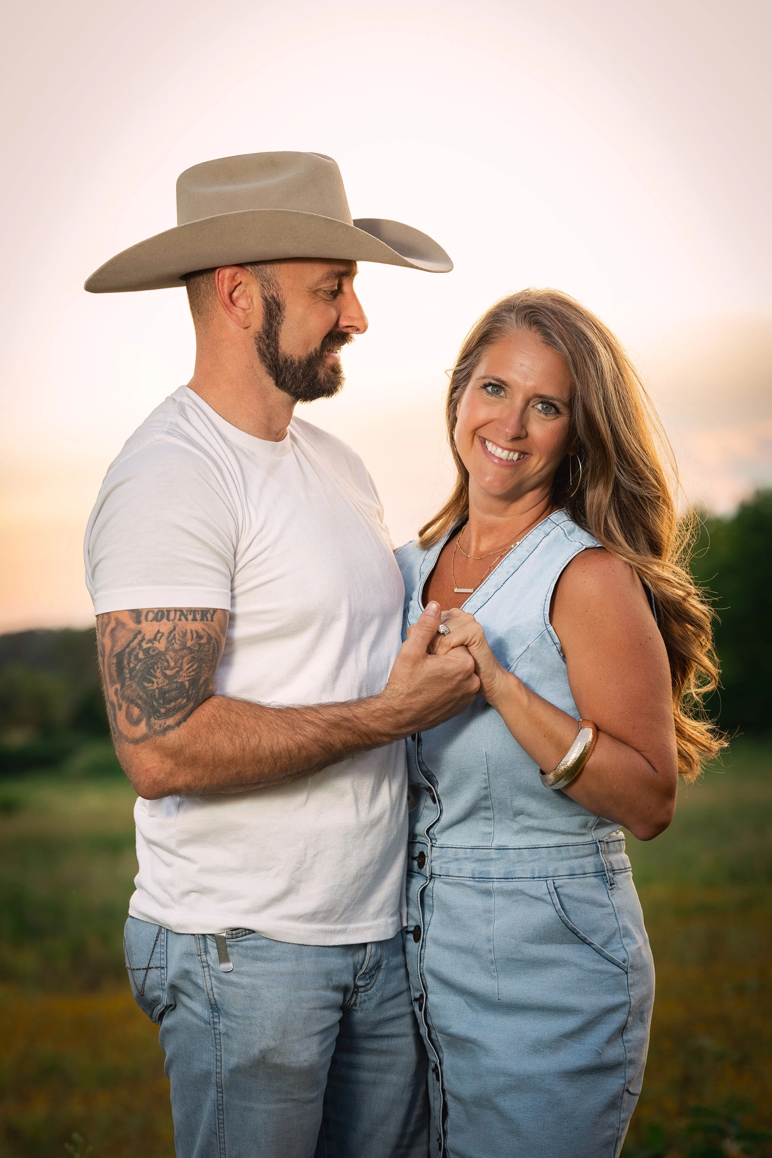 A smiling woman and a man with a beard and cowboy hat holding hands outdoors during sunset.