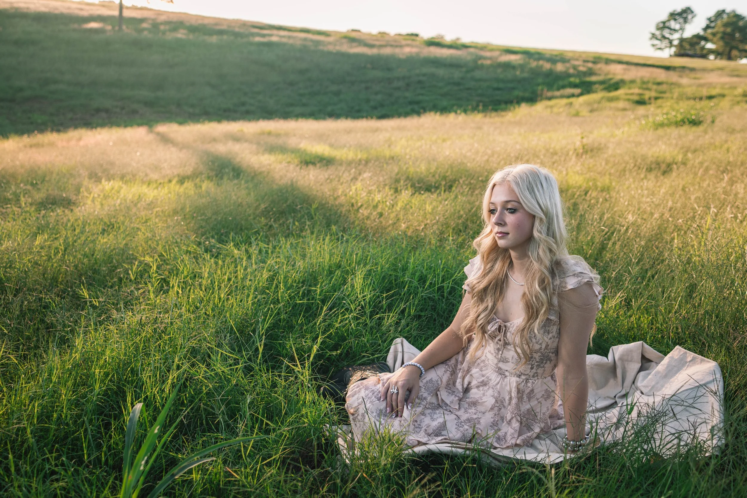 A young woman with long blonde hair sitting in a grassy field during sunset, wearing a light-colored dress and looking contemplative.