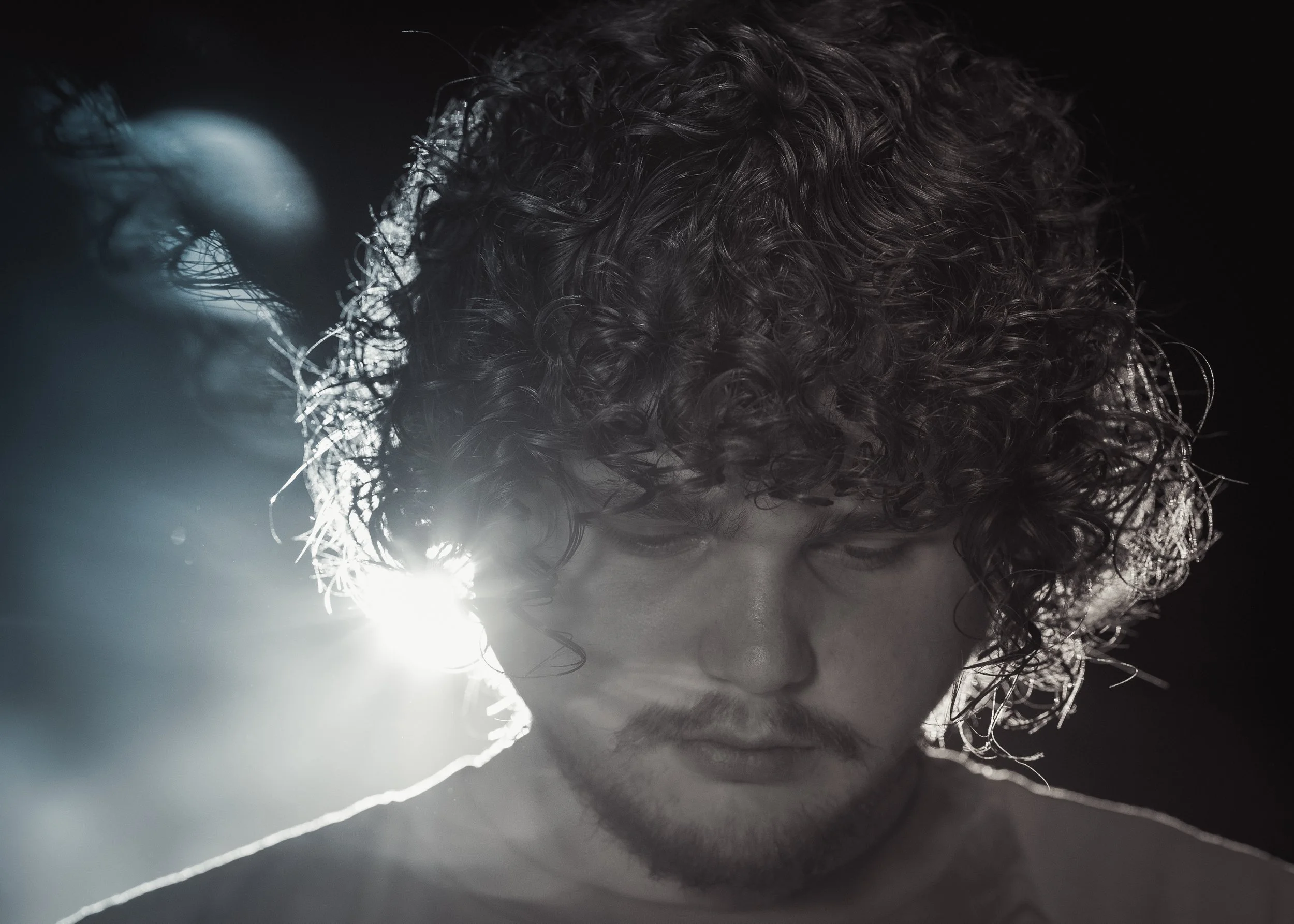 Black and white close-up photo of a young man with curly hair looking downward, with sunlight reflecting behind his head.