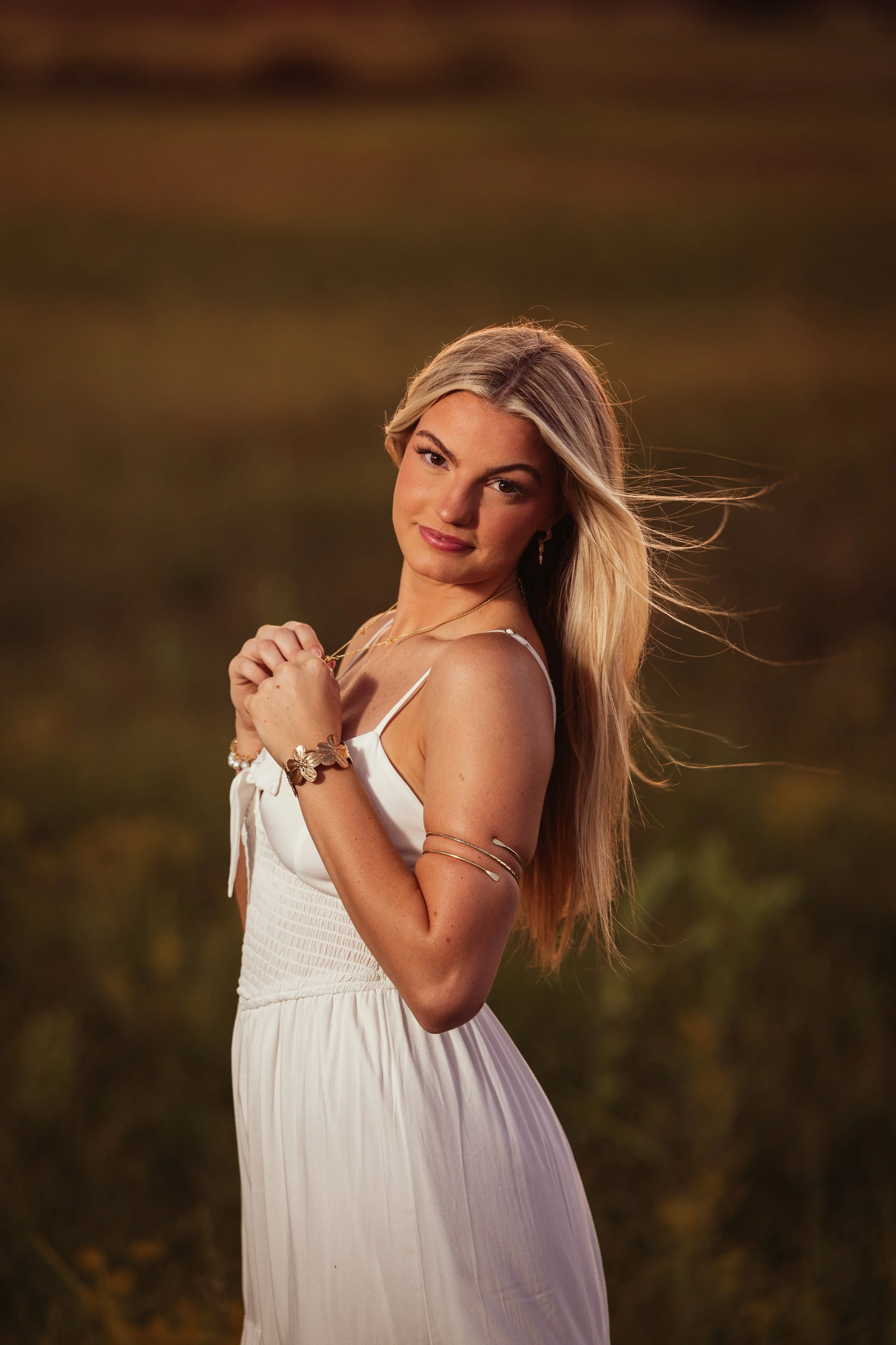 A young woman with long blonde hair, wearing a white dress and jewelry, standing outdoors in a natural setting during sunset.