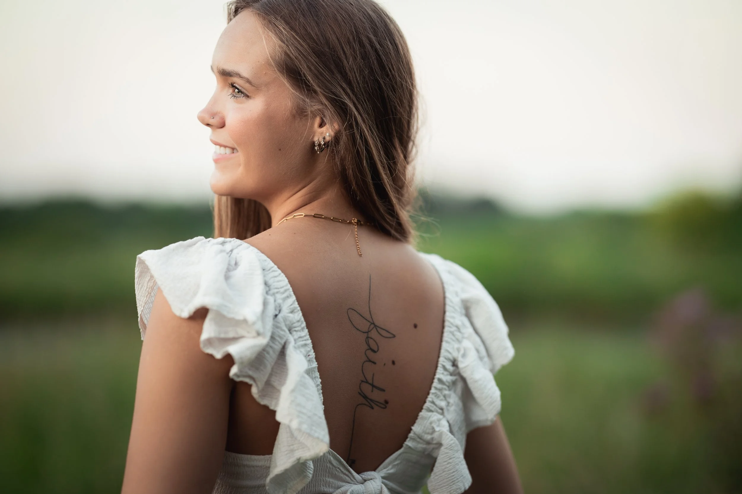 Young woman with long brown hair, wearing a white dress with ruffled sleeves, looking to the left with a smile, outdoor green landscape in the background, showing a tattoo on her back that says "faith" in cursive.