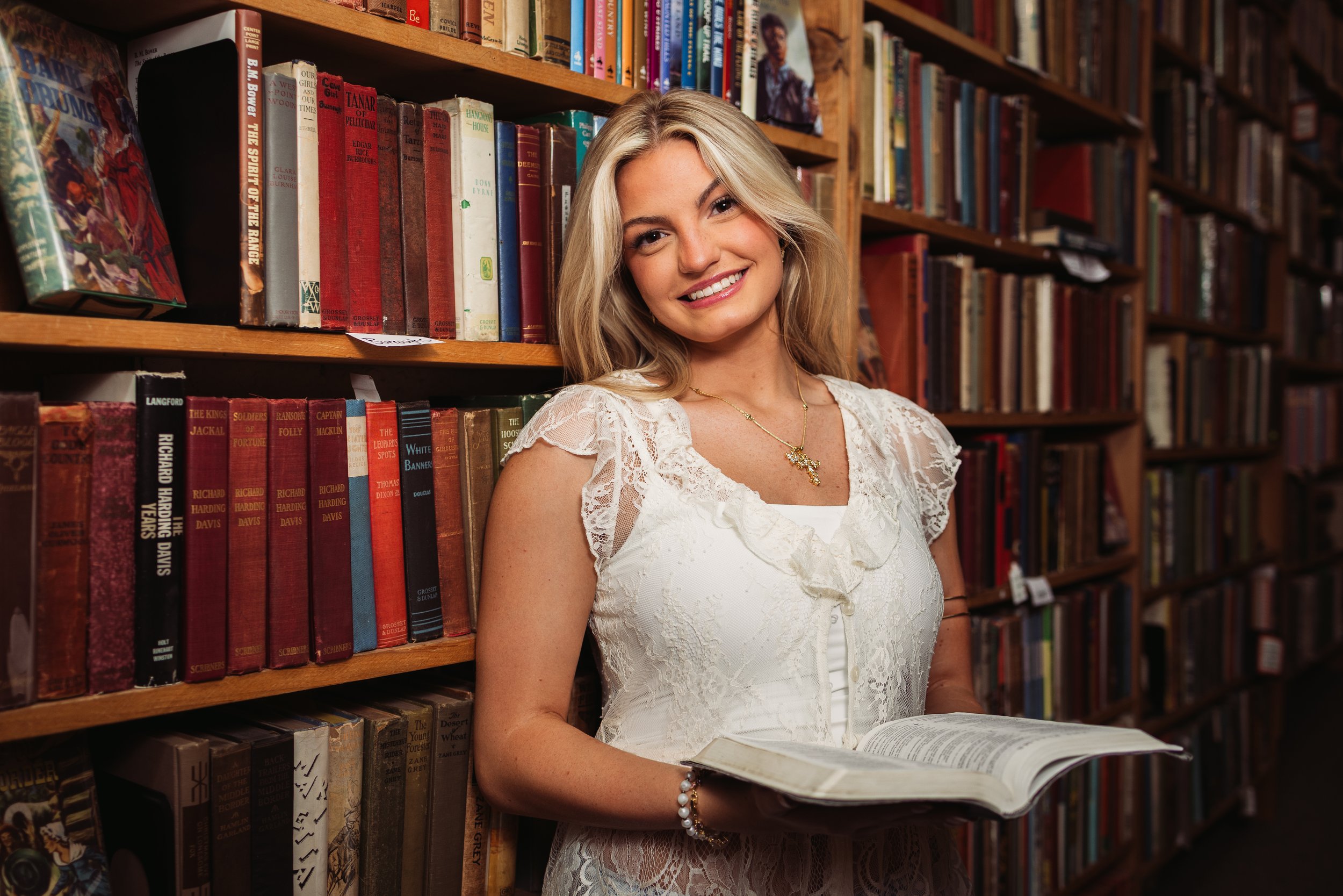 A young woman with long blonde hair smiling and holding an open book in a library with wooden bookshelves filled with colorful books. Senior Class of 2027 portrait photographer best photographer in northwest arkansas