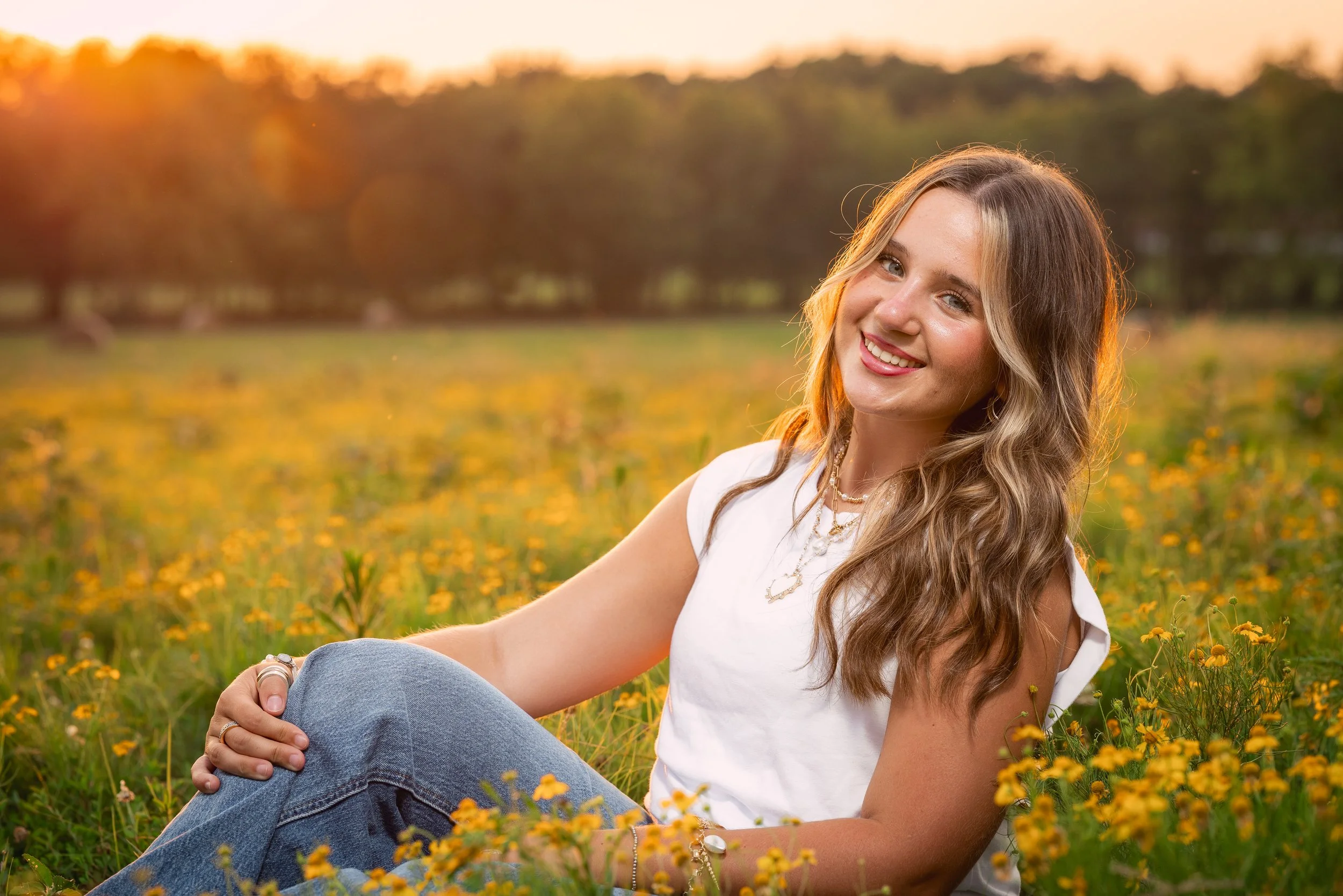 A smiling young woman with long wavy hair sitting in a field of yellow flowers at sunset.