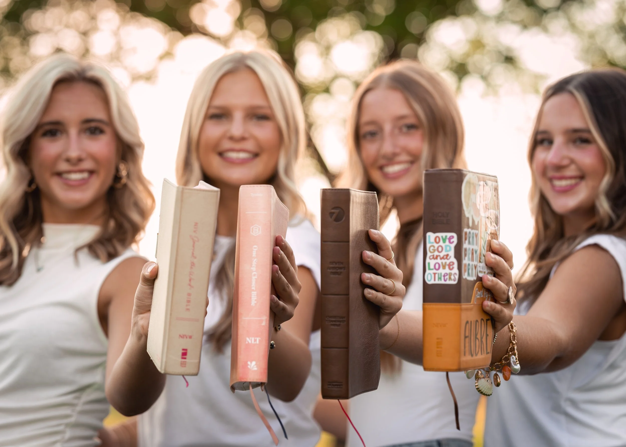 Four young women smiling and holding up books outdoors with trees and sunlight in the background.