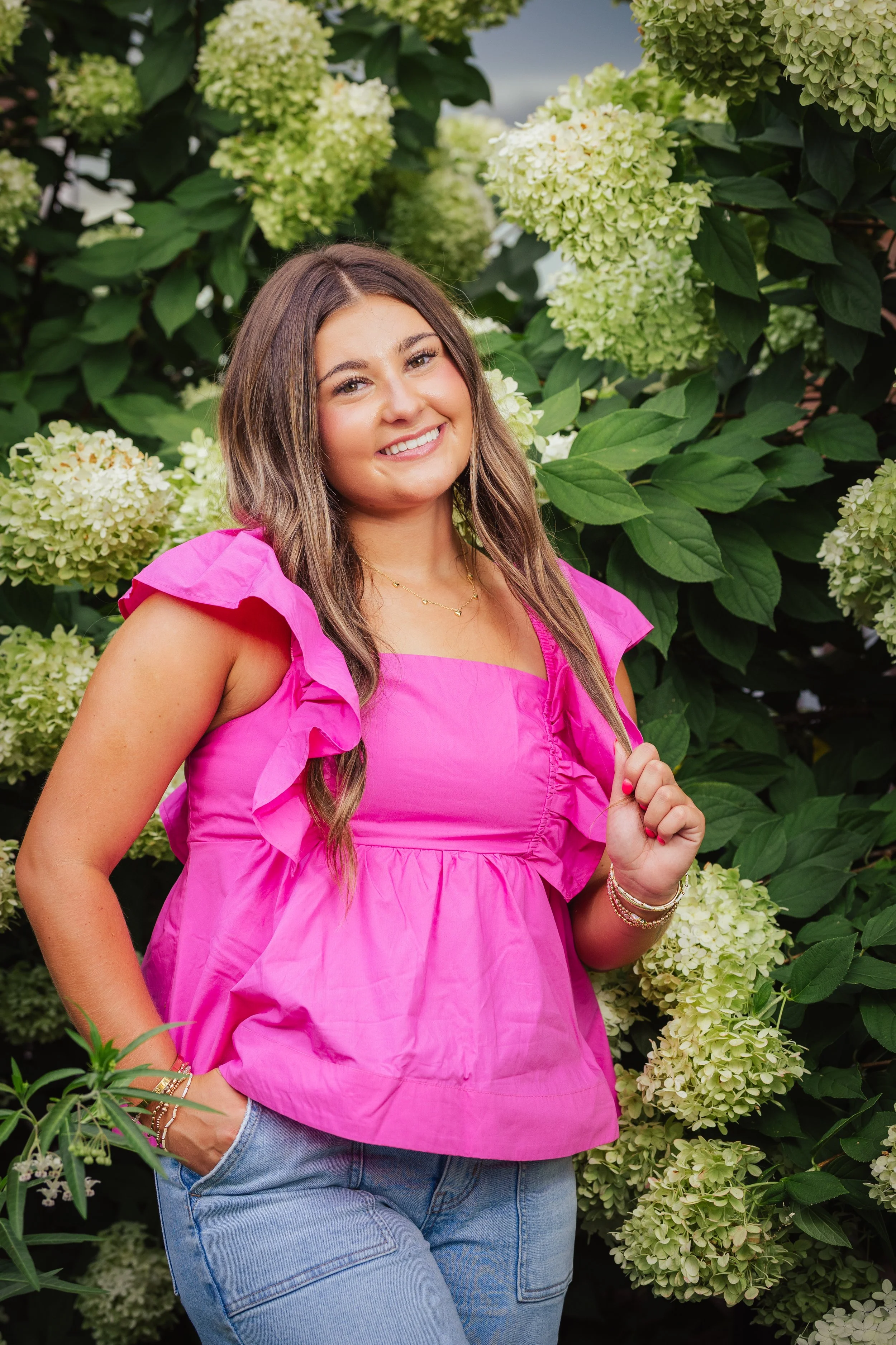 A woman with long brown hair smiling, wearing a bright pink top with ruffled sleeves, standing in front of green hydrangea bushes.