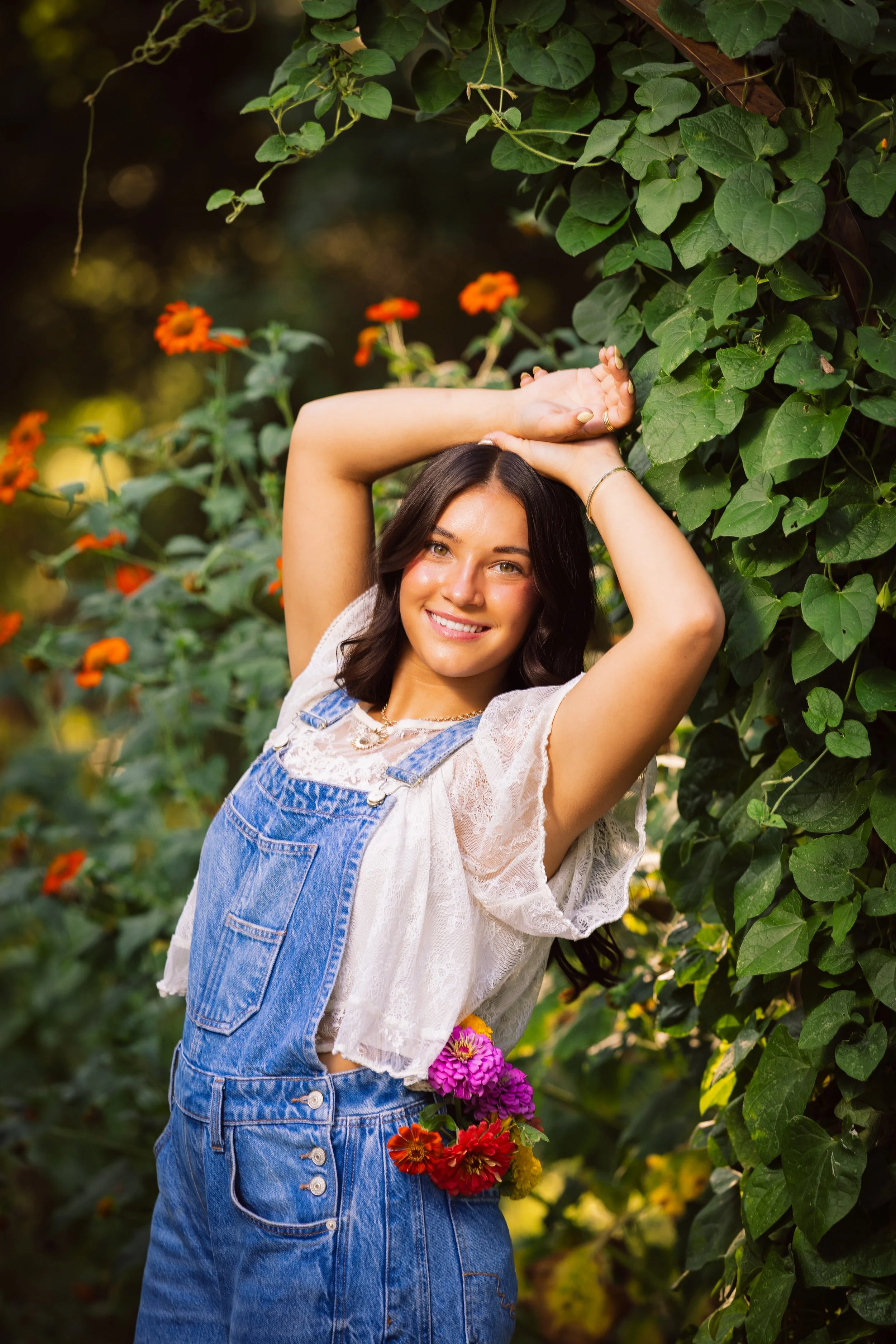 A woman with long dark hair smiling, wearing a white lace top and denim overalls, standing amidst green foliage and colorful flowers, with her arms raised above her head.