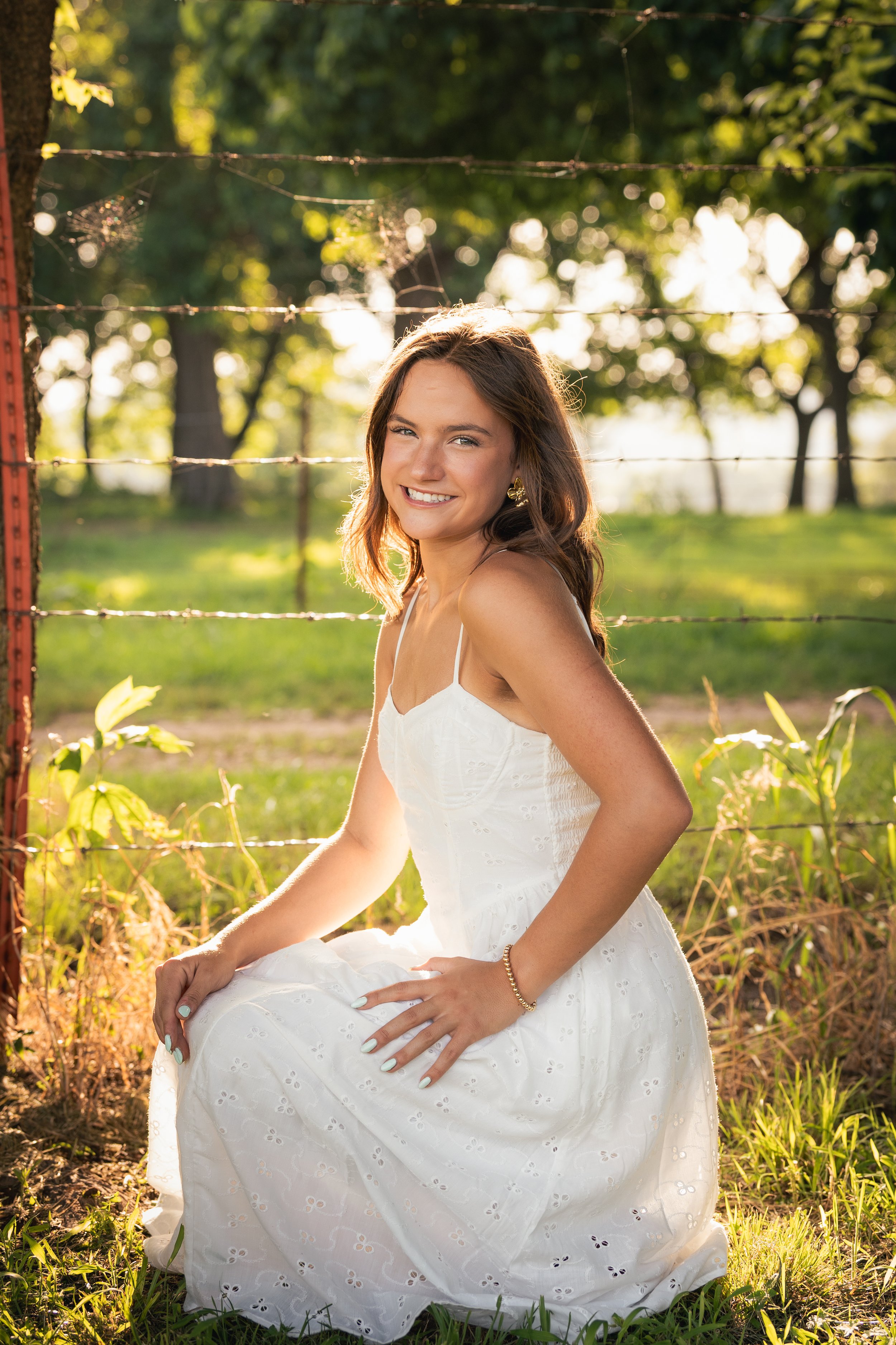 A young woman in a white dress sitting outdoors near a barbed wire fence with green trees in the background, smiling and illuminated by warm sunlight.