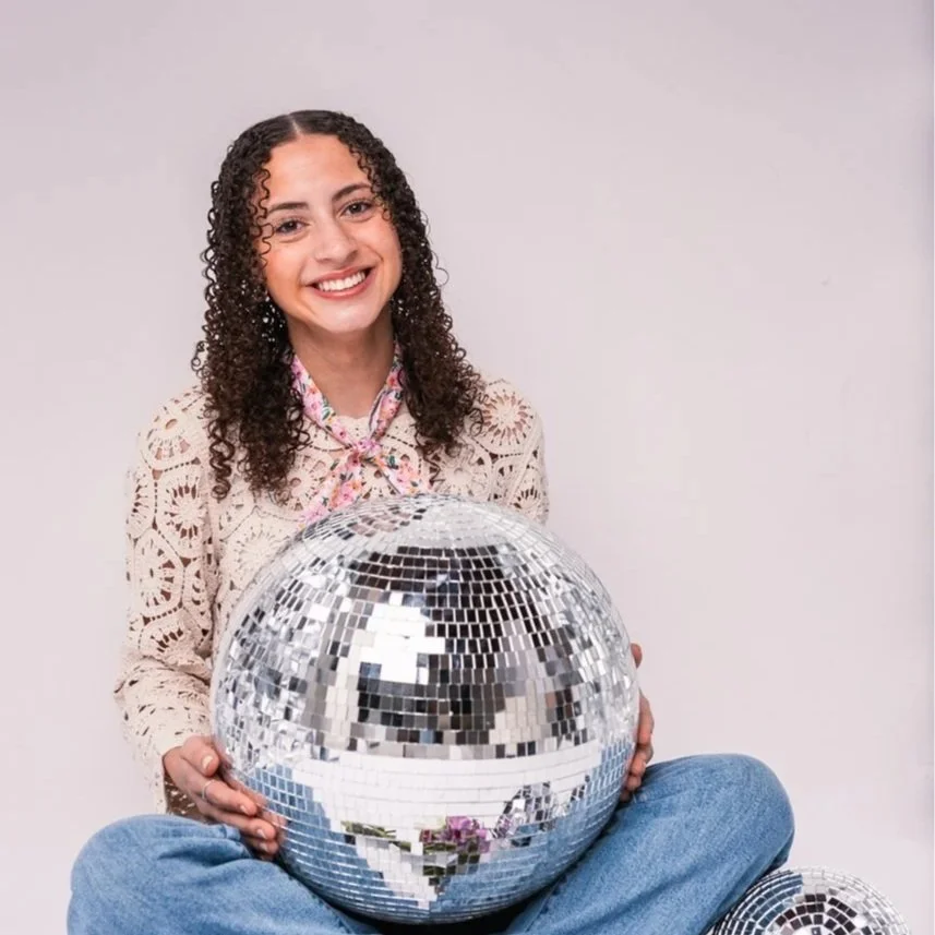 A young woman with curly hair smiling and sitting cross-legged on the floor, holding a large disco ball in her lap.
