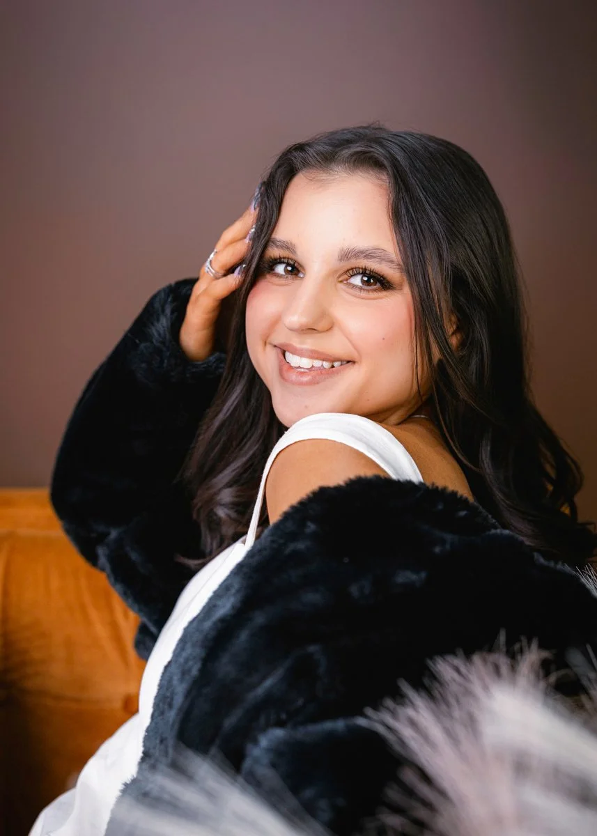 A young woman smiling, with long dark hair, sitting on a wooden chair, wearing a white top and a black fuzzy jacket, with one hand touching her hair, against a brown background.