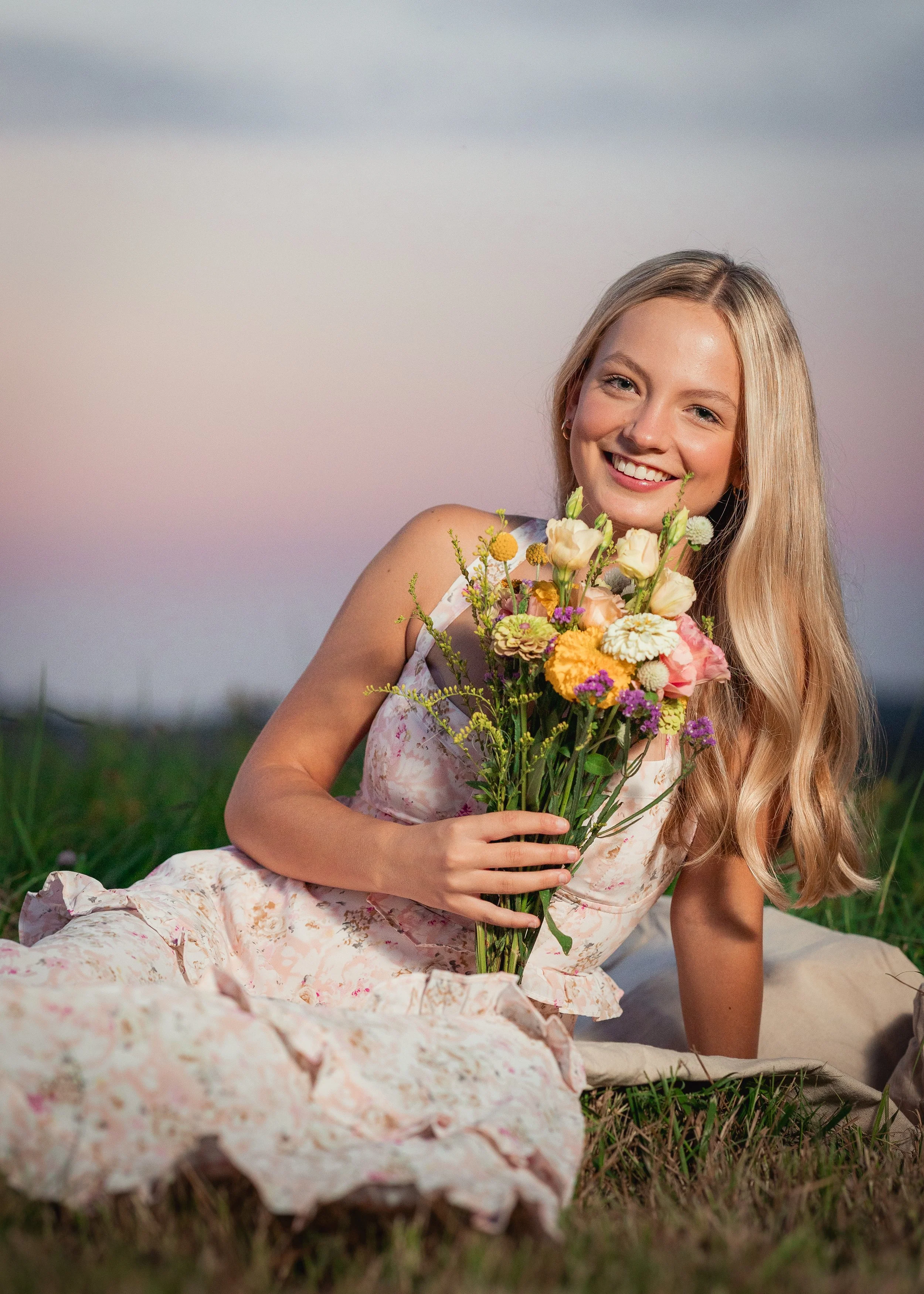 A young woman with long blonde hair lying on the grass, holding a bouquet of colorful flowers, smiling, with a pastel-colored sky in the background.