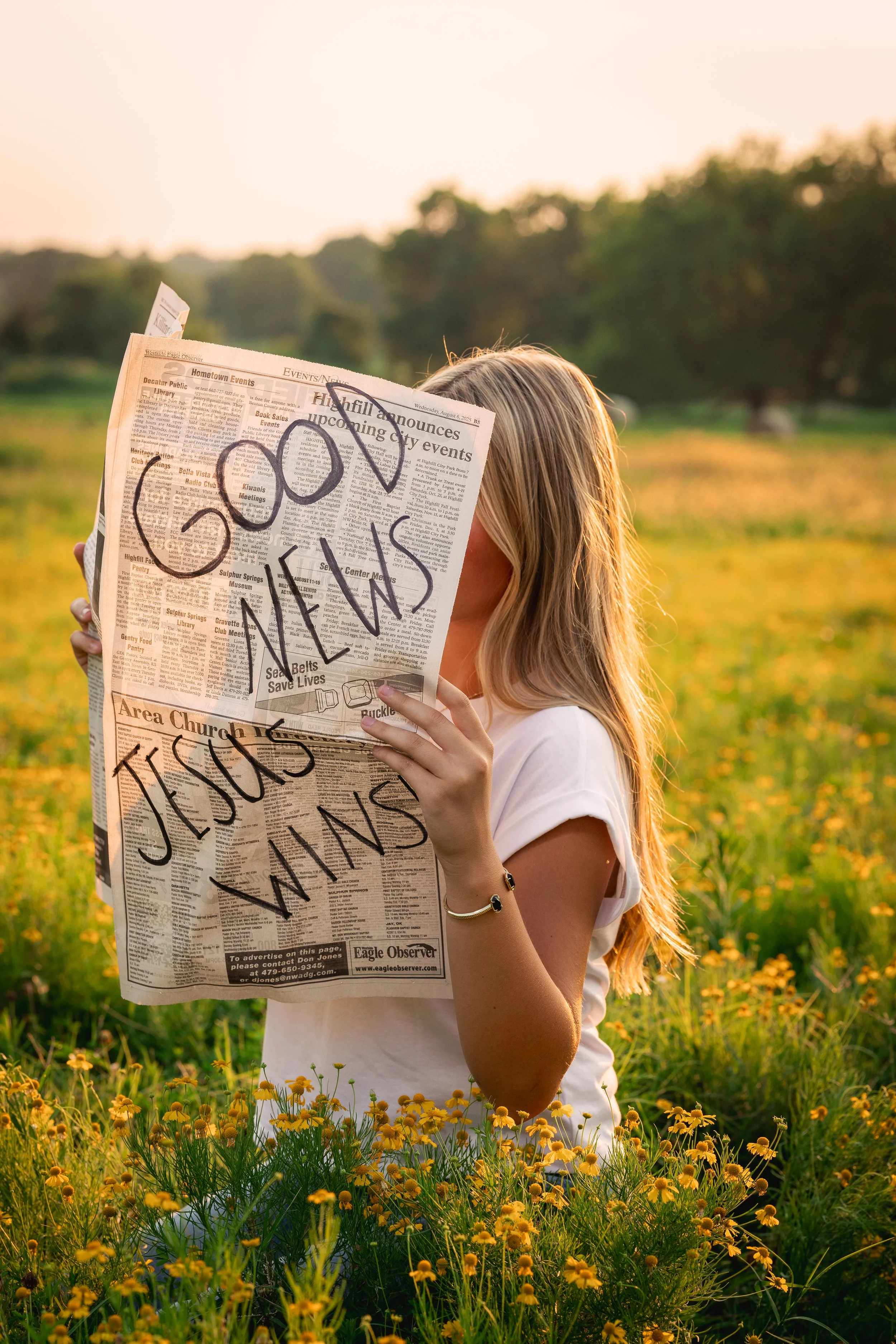 A woman with blonde hair holding a newspaper in a field of yellow flowers at sunset, with handwritten message on the newspaper that reads 'Good vibes, Jesus wins'.
