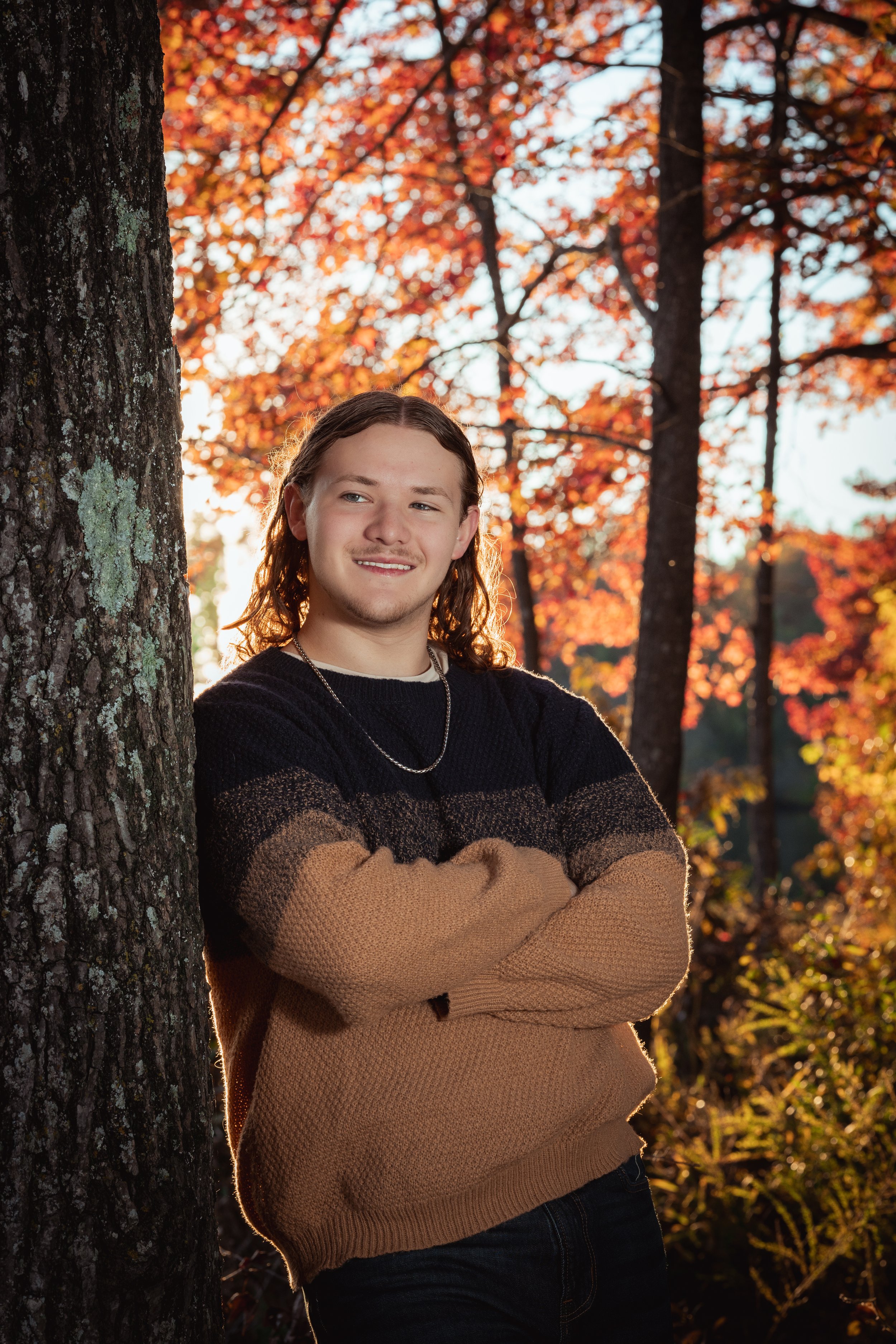 A young man with long, wavy hair leaning against a tree in a forest with autumn foliage, arms crossed, smiling softly, during sunset.