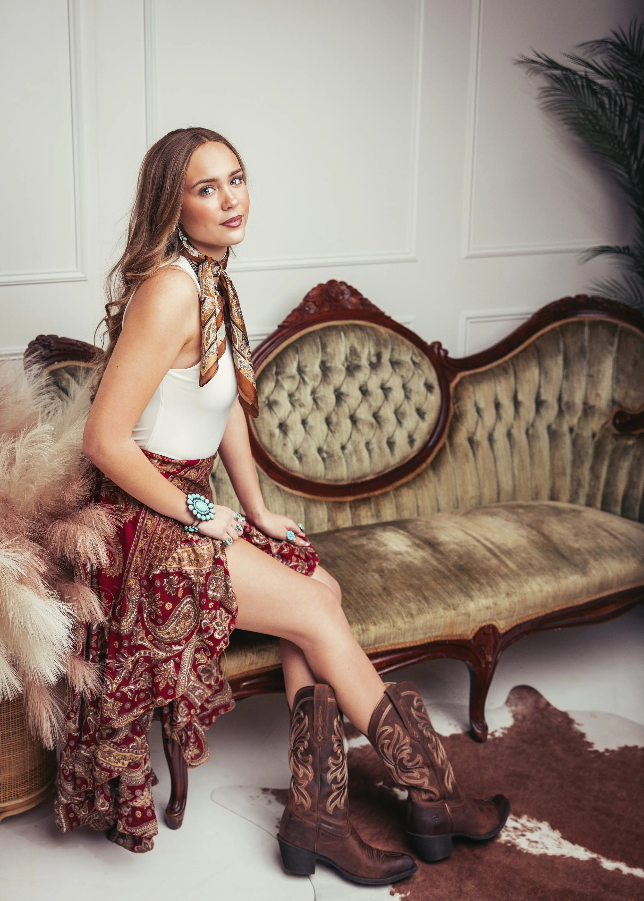 Woman sitting on a vintage sofa in a room with white paneled walls, wearing cowboy boots, a white top, a patterned skirt, and a scarf around her neck, with jewelry and a feathered accessory.