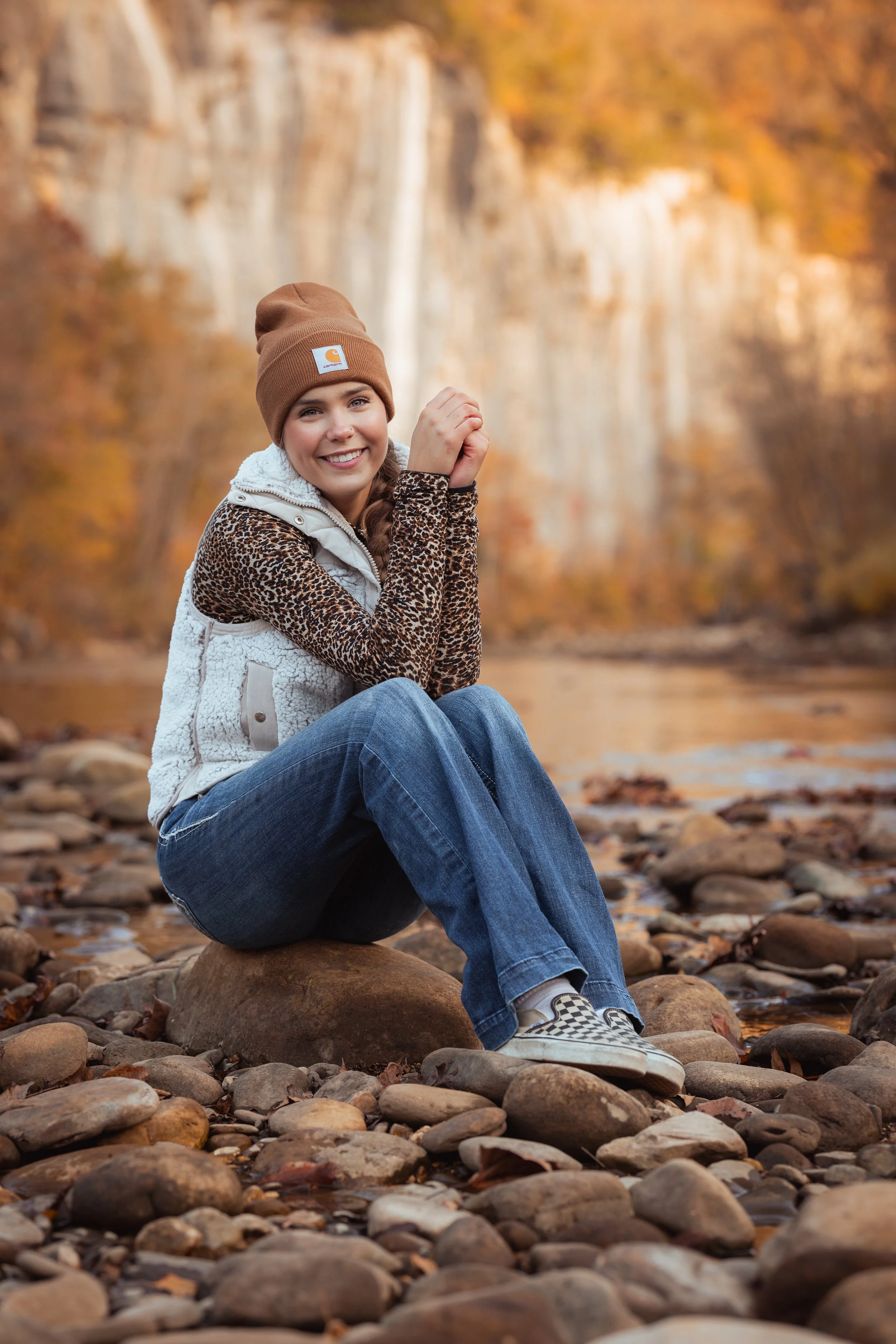 Young woman sitting on rocks by a river with autumn trees in the background, smiling at the camera, wearing a brown beanie, leopard print long sleeve shirt, white vest, blue jeans, and checkered slip-on shoes.