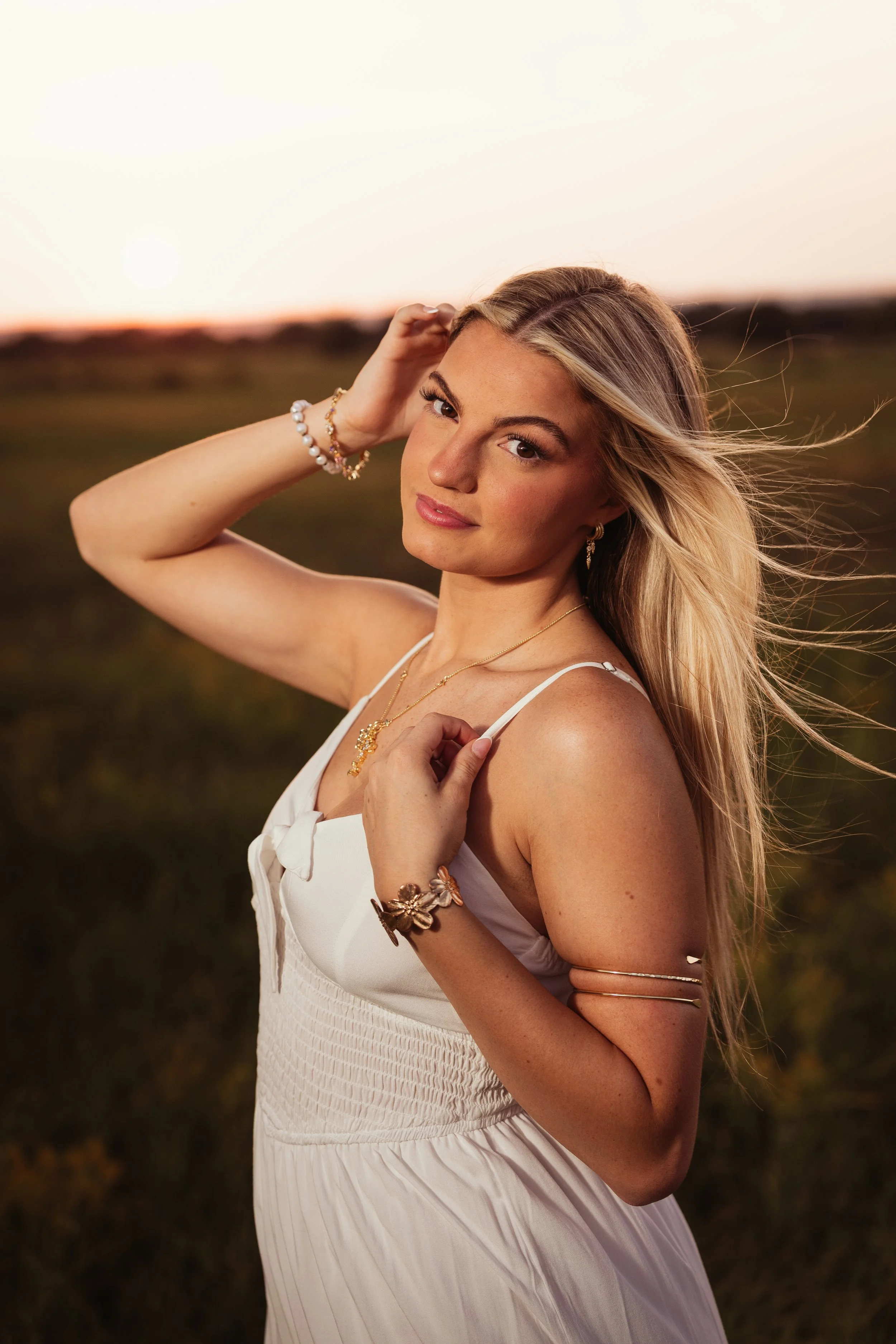 A young woman with long blonde hair is posing outdoors during sunset, wearing a white dress and jewelry, and looking at the camera.