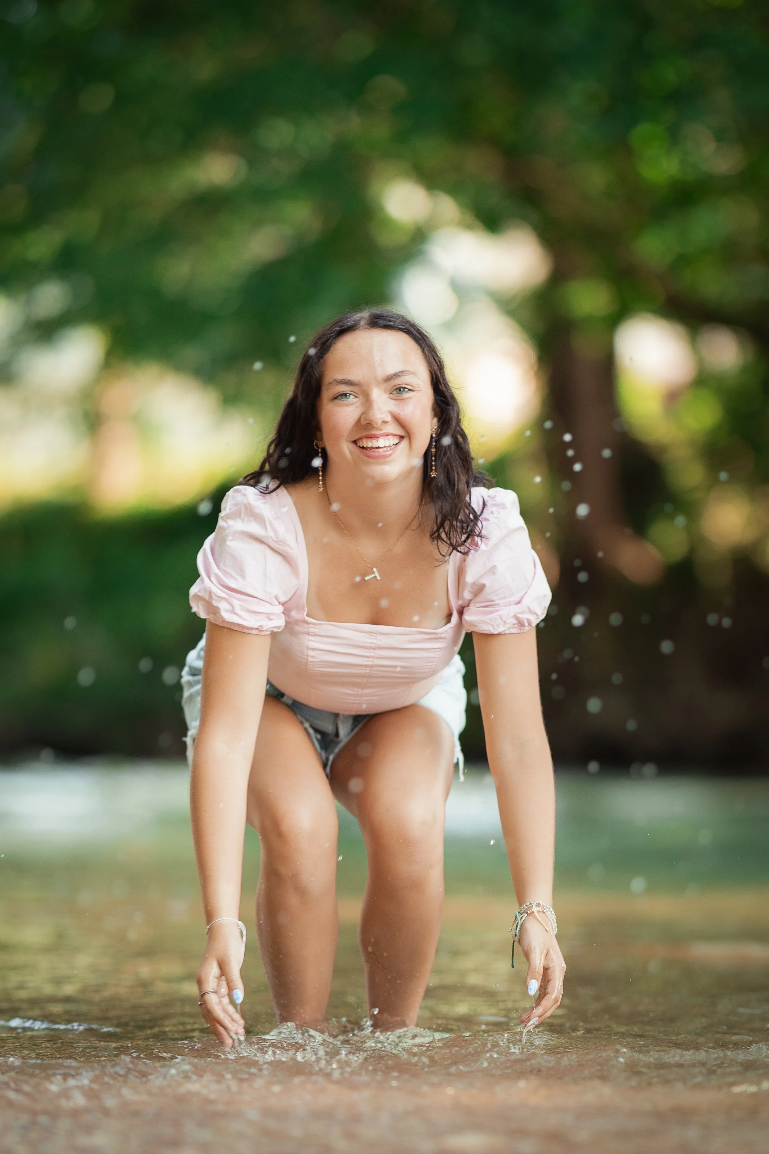 A woman crouching in a shallow stream, smiling, with trees and greenery in the background.