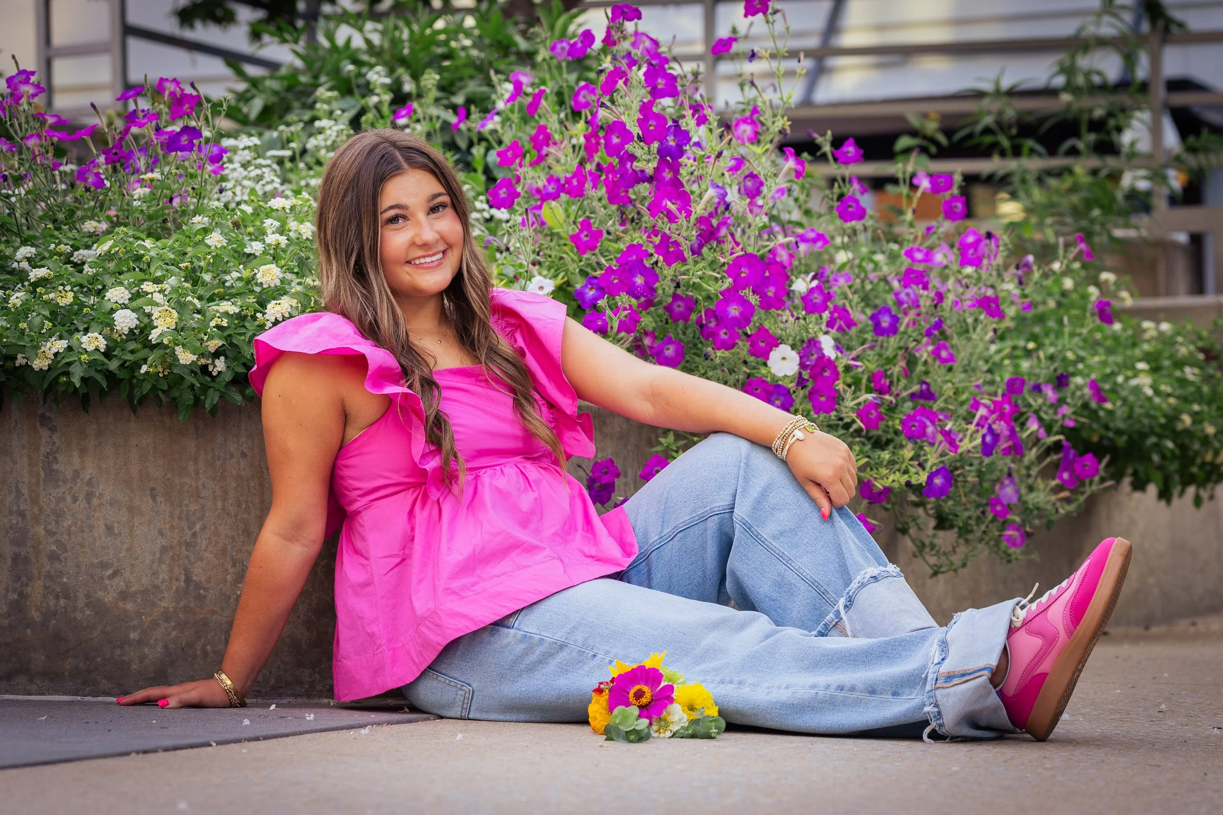 A young woman sitting on the ground near a flower bed, smiling, wearing a bright pink top and jeans, with colorful flowers on the ground in front of her.