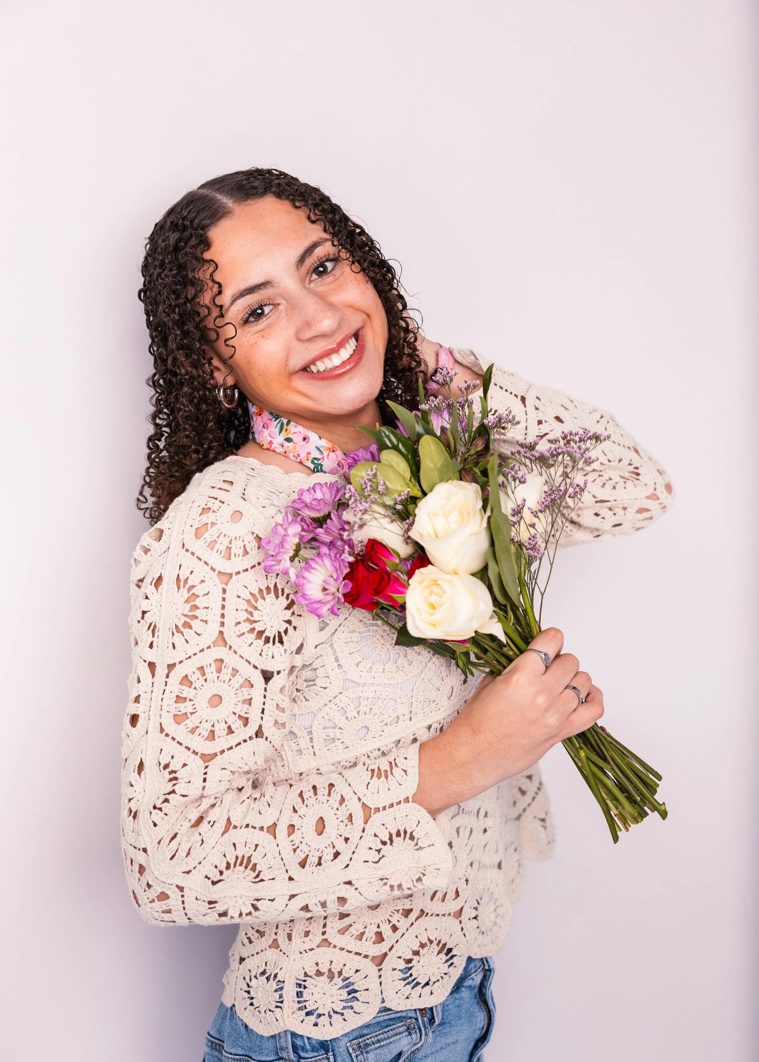 A woman with curly hair smiling and holding a bouquet of mixed flowers, standing against a plain white background.
