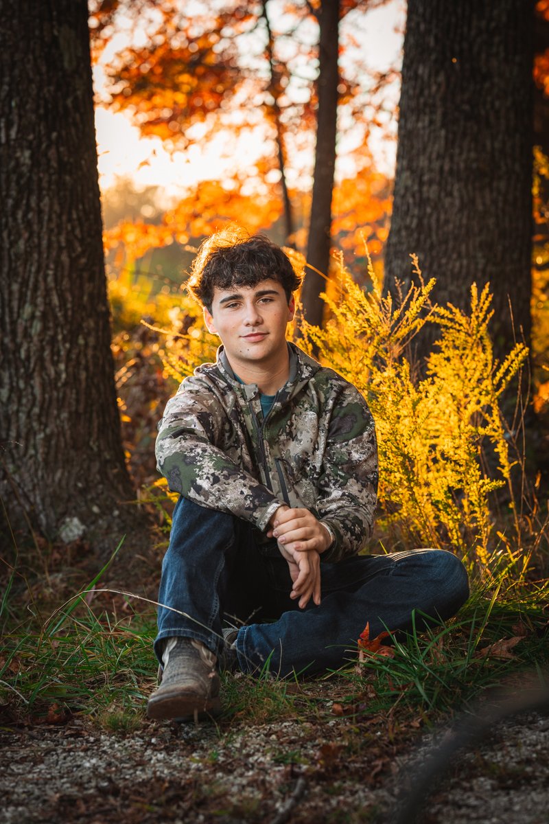 A young man sitting on the ground with crossed legs, wearing a camouflage jacket, in a forest with autumn foliage, illuminated by a warm sunset.