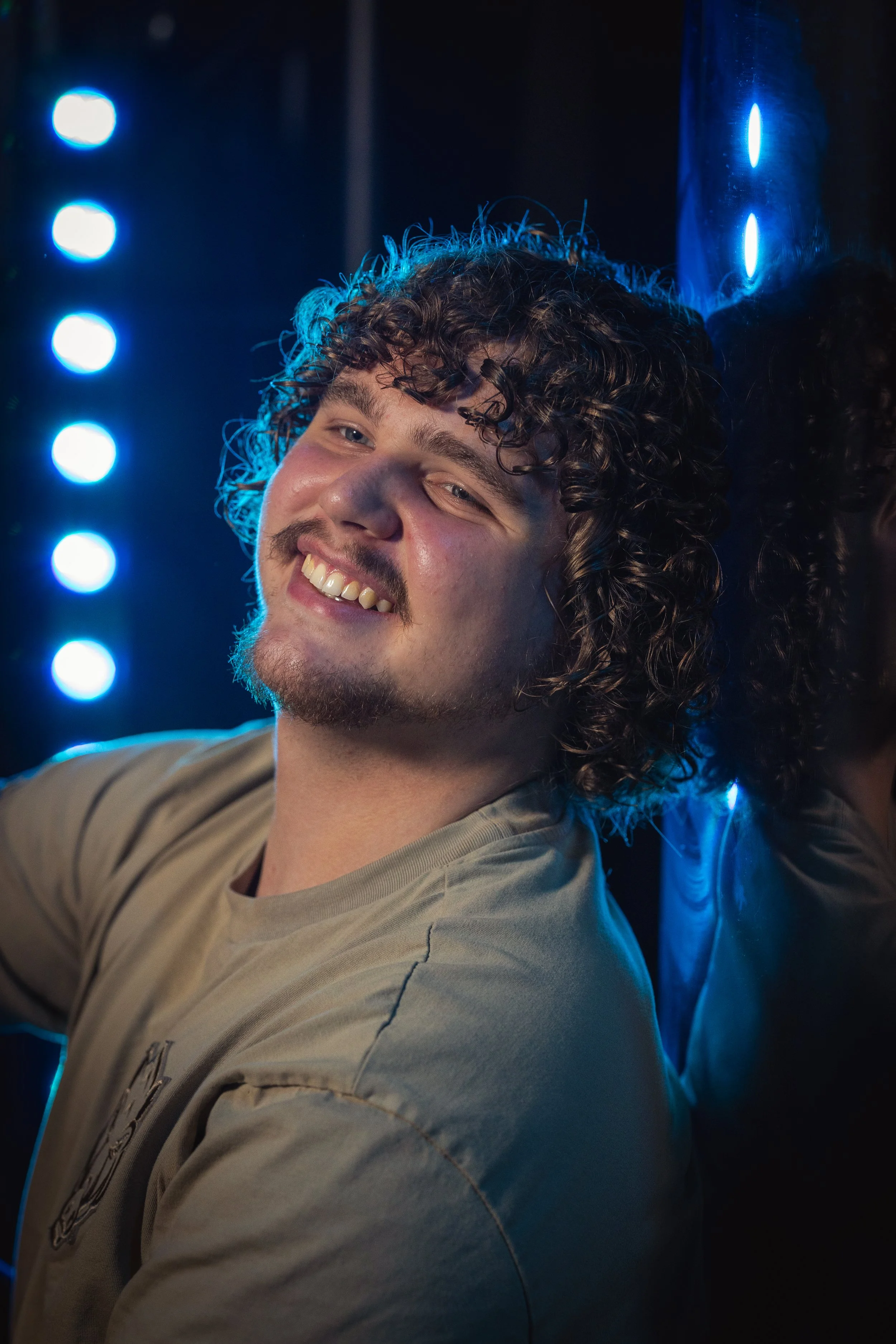 A young man with curly hair and a beard smiling while leaning against a reflective surface with blue LED lights in the background.