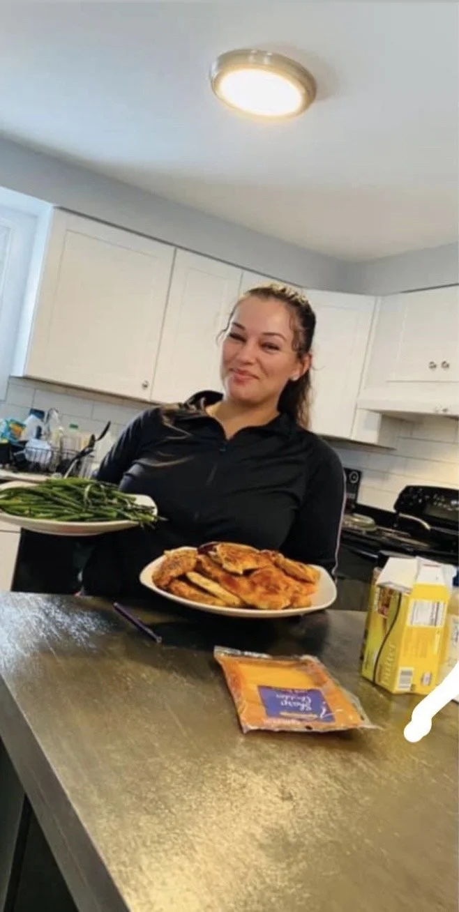 A woman in a black shirt standing in a kitchen, smiling at the camera, with two plates of food on the counter in front of her, including green beans and fried chicken, along with a packet of dipping sauce and a container of juice.