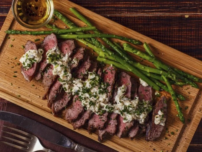 Sliced steak topped with herb sauce, served with asparagus on a wooden platter.