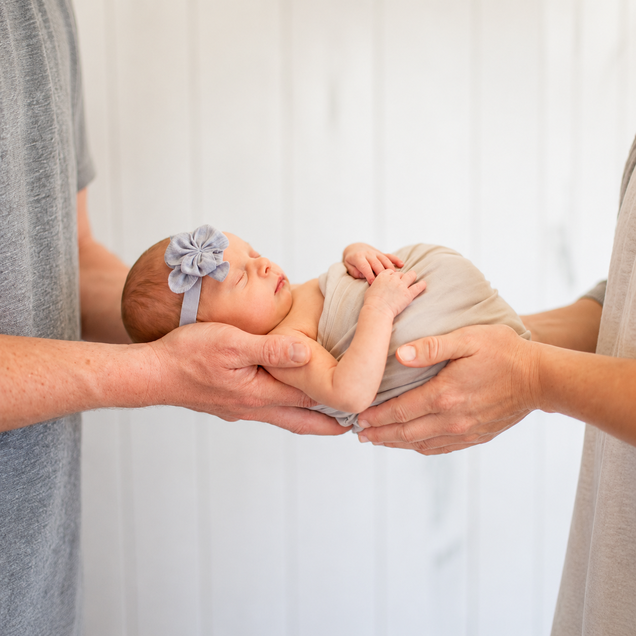 A newborn baby girl with a blue bow headband is being gently held in the hands of two adults against a white background.