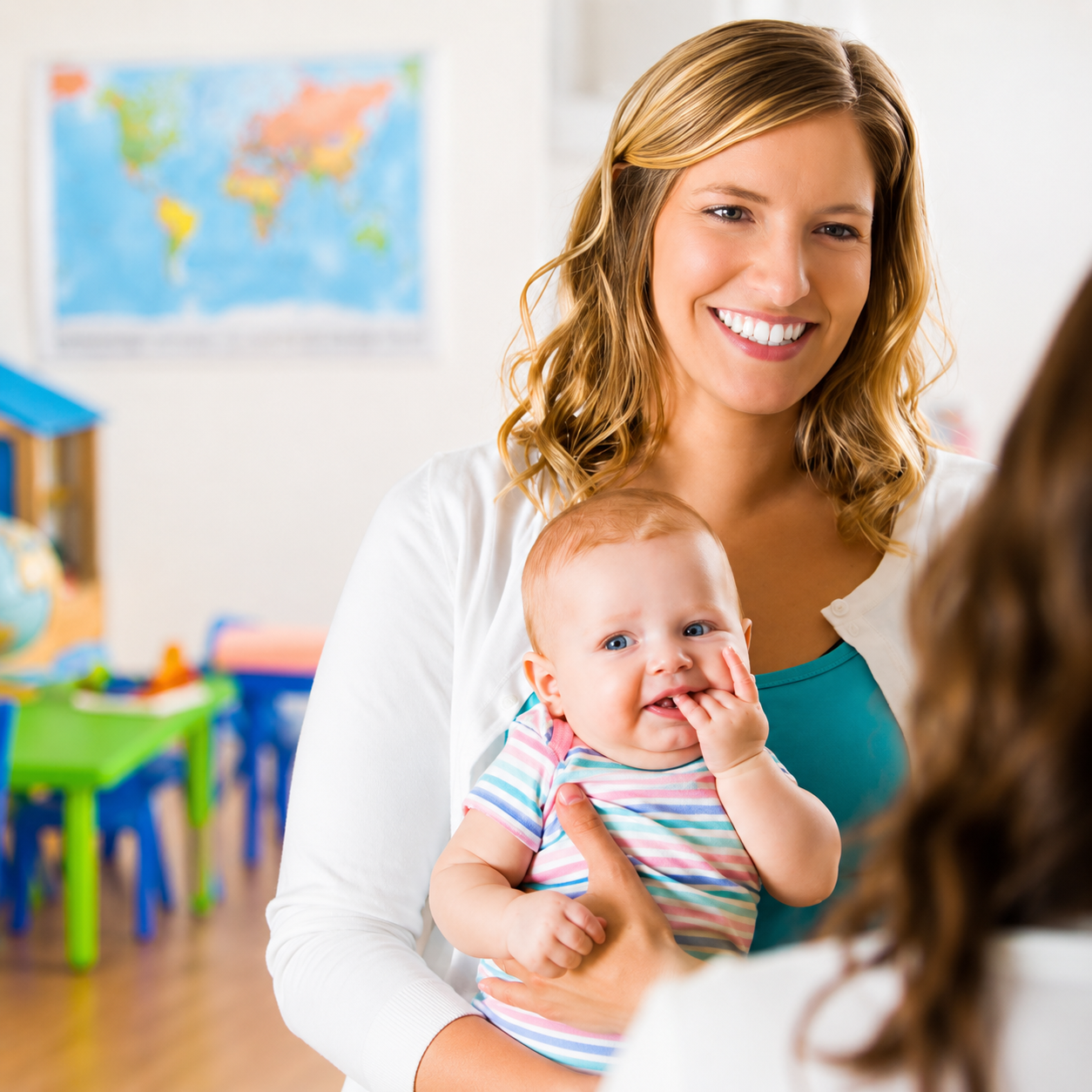 A smiling woman holding a baby in a classroom with colorful furniture and a world map on the wall.