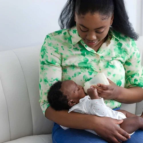 Mother gently bottle-feeding her baby while sitting on a white sofa at home.