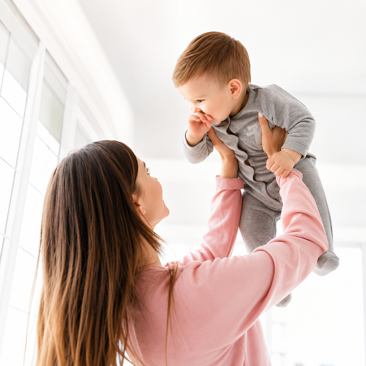 A woman with long brown hair holding a young boy in gray clothes high above her head, both smiling and looking at each other inside a bright room with large windows.