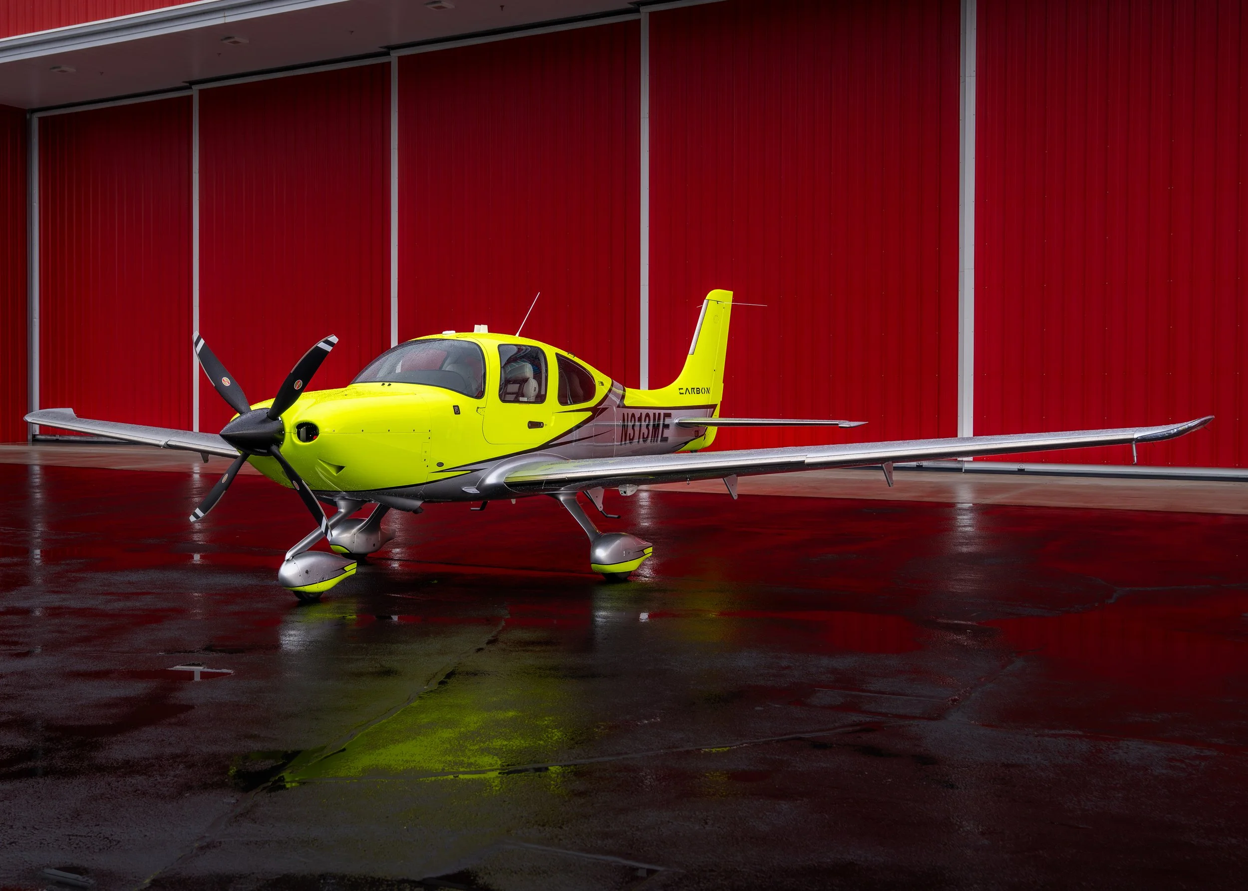 Bright yellow light aircraft with black nose and propellers, parked on a wet concrete floor in front of a red corrugated metal wall.