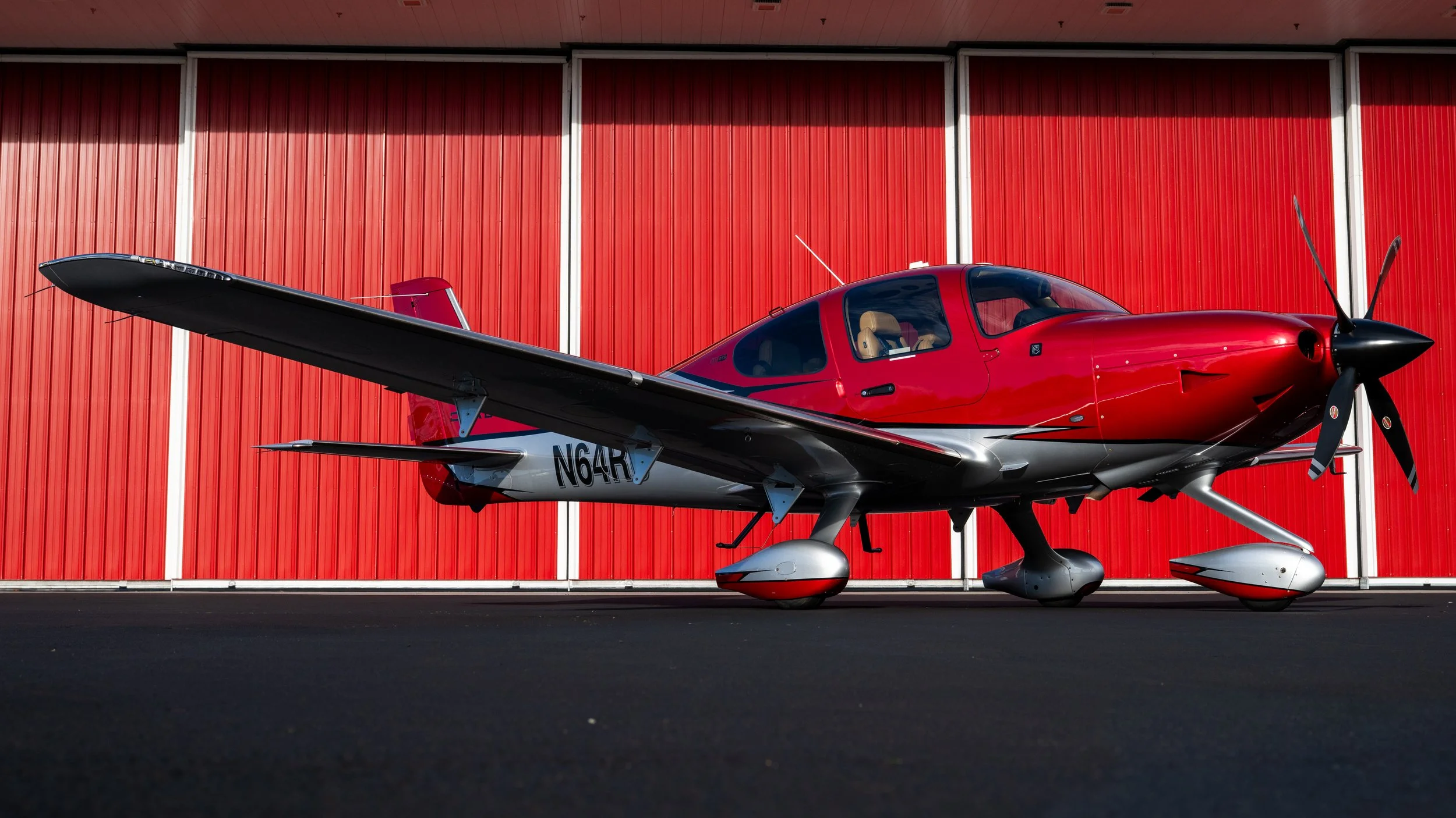 A red and black small airplane parked on tarmac in front of a red hangar.