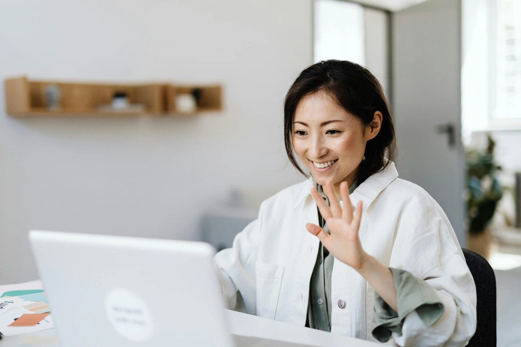A woman with short dark hair smiling and waving at her laptop in a bright, modern room.