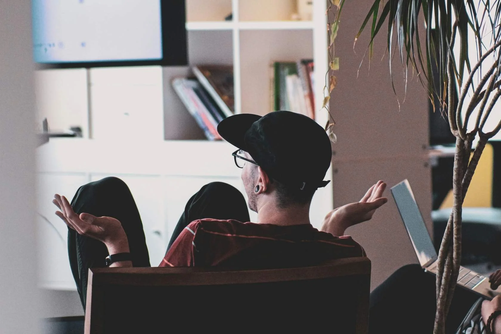 A person with black hair, wearing glasses and a black cap, sitting on a chair indoors with arms raised and palms facing upward, in front of a laptop, surrounded by bookshelves and a large plant.
