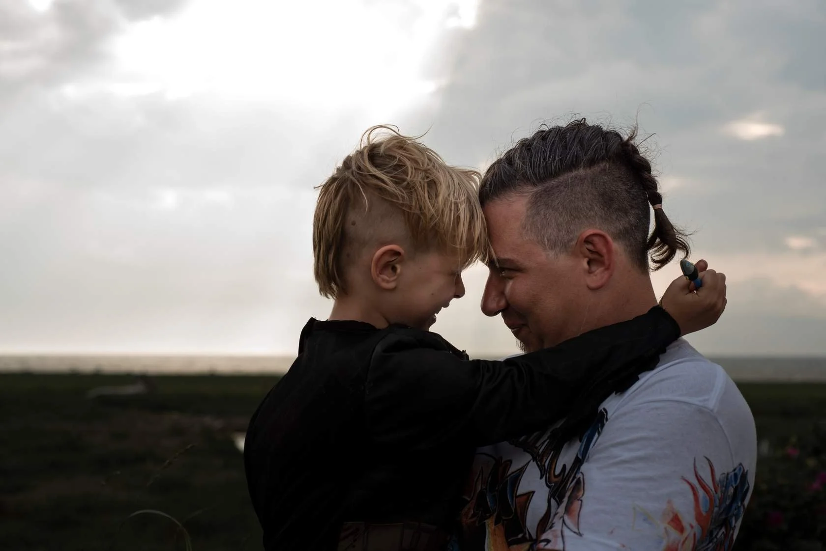A young boy and an adult man with styled hair are touching foreheads and smiling at each other outdoors during cloudy weather.
