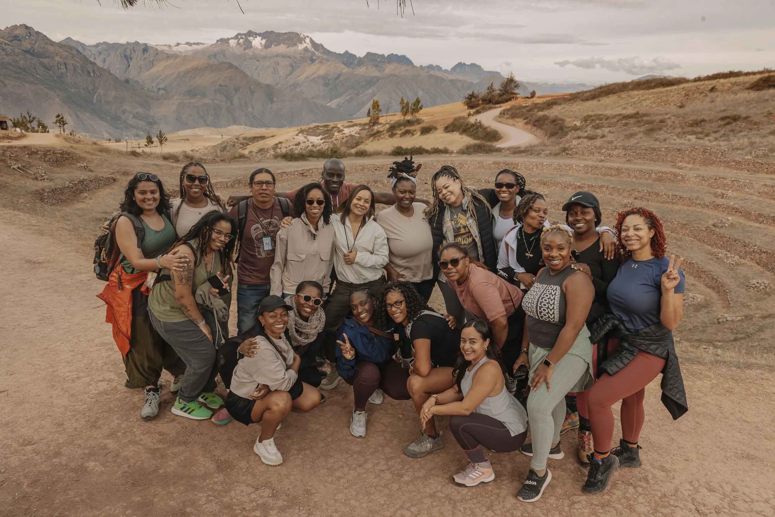 Group of women and a man smiling and posing outdoors on a dirt path with mountainous landscape in the background.