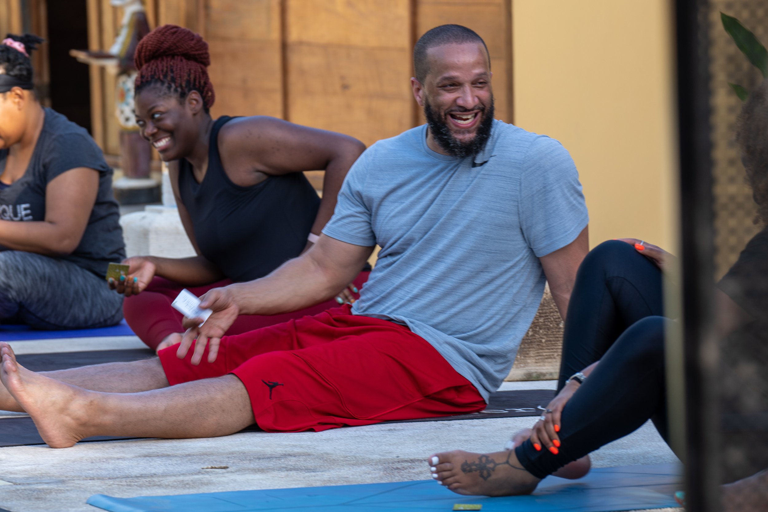 Group of people sitting on yoga mats, smiling, and engaging in a yoga or relaxation class in a studio with wooden walls.
