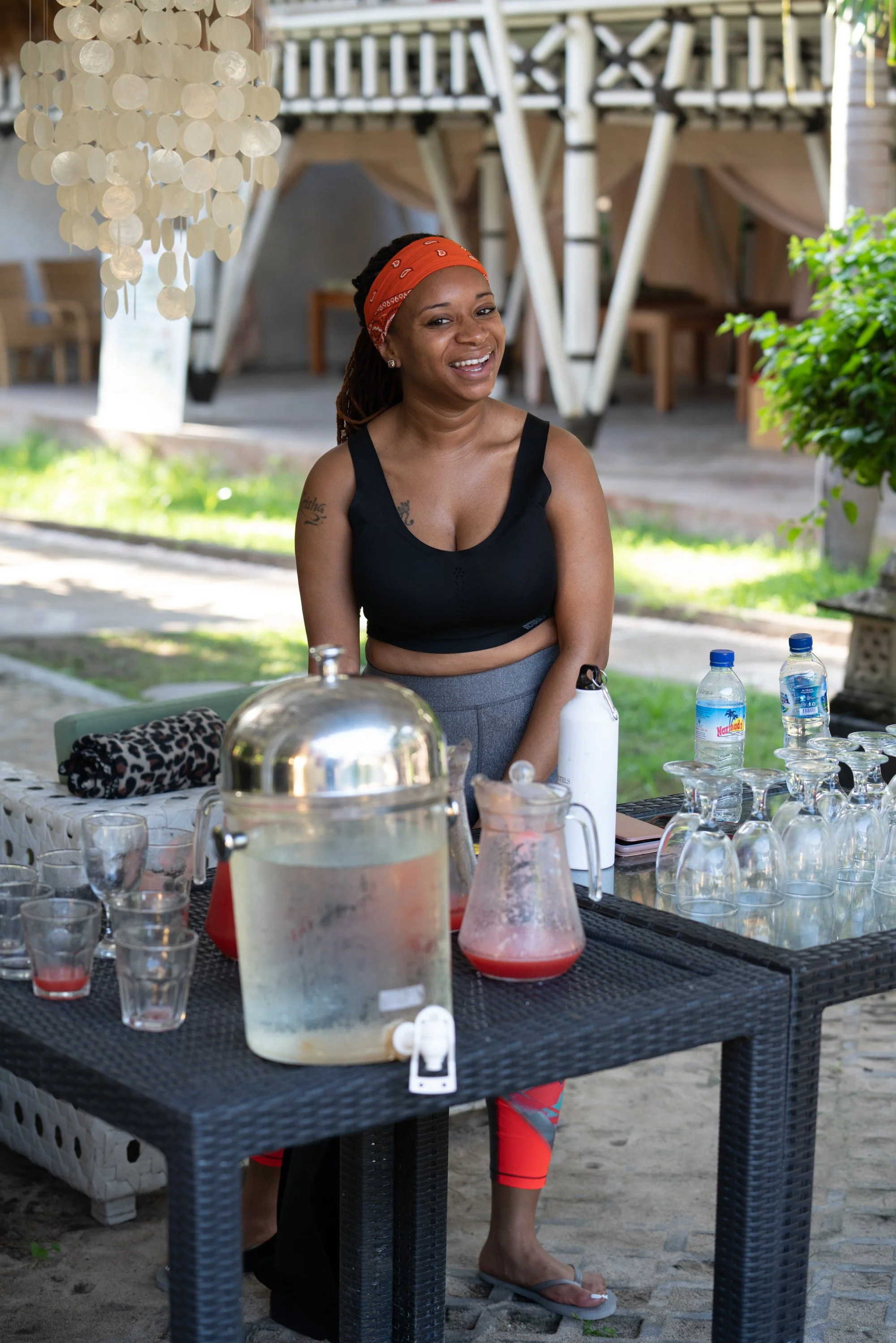A woman with a red bandana smiling standing behind a black outdoor table with drinks, water bottles, glasses, and a pitcher, in a sunny outdoor setting with greenery and wooden structures.