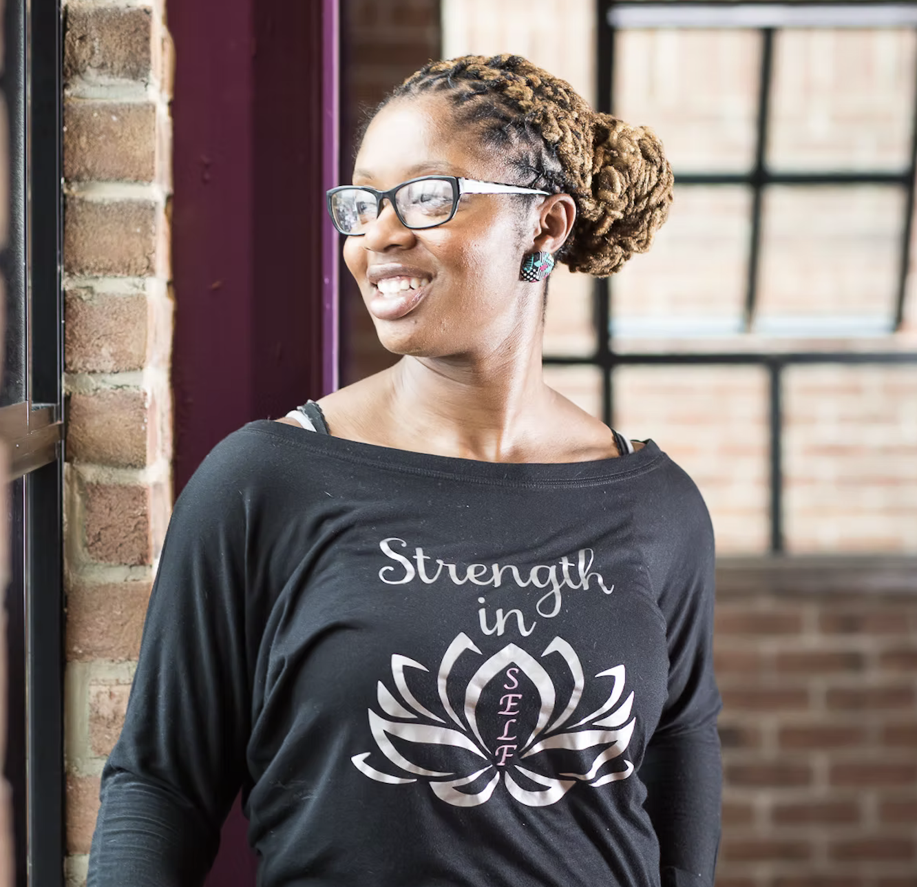 A woman with glasses and styled dreadlocks smiling and looking to the side. She is wearing a black shirt with the words 'Strength in Self' and a lotus flower design.